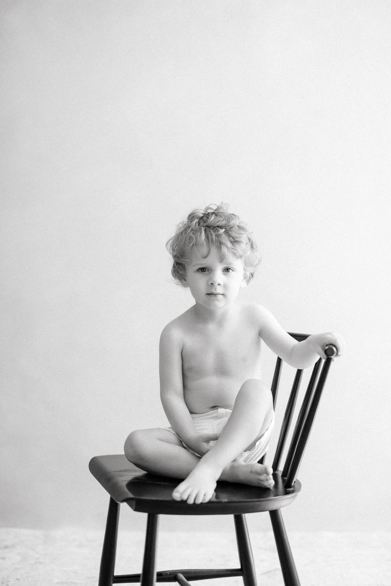 Portrait of a toddler sitting on a wooden chair during a family newborn photography session.