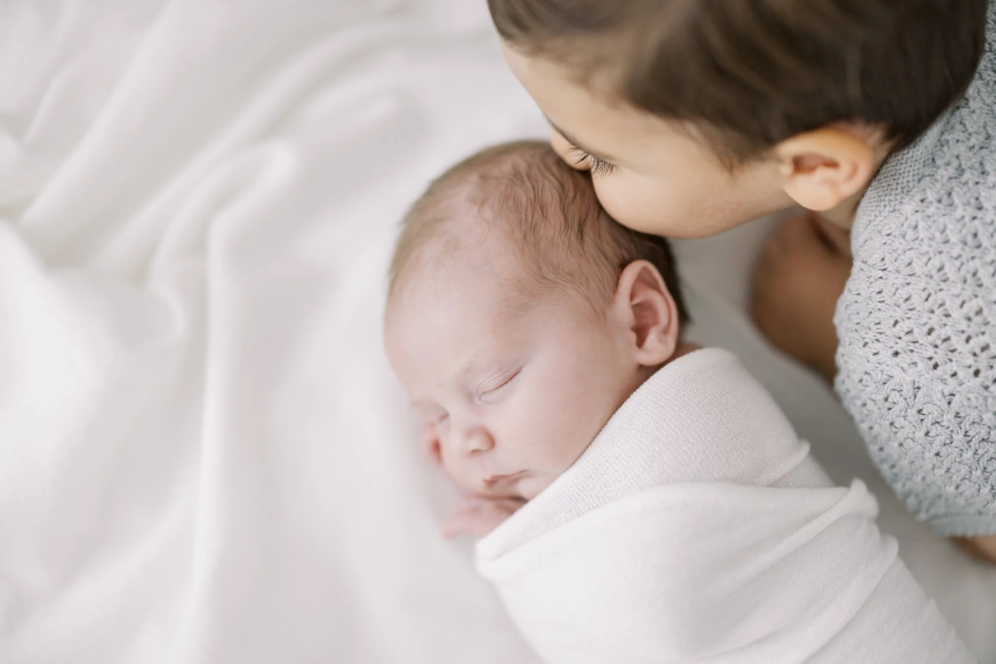 Older brother kissing his sleeping newborn sibling during a family newborn photography session.