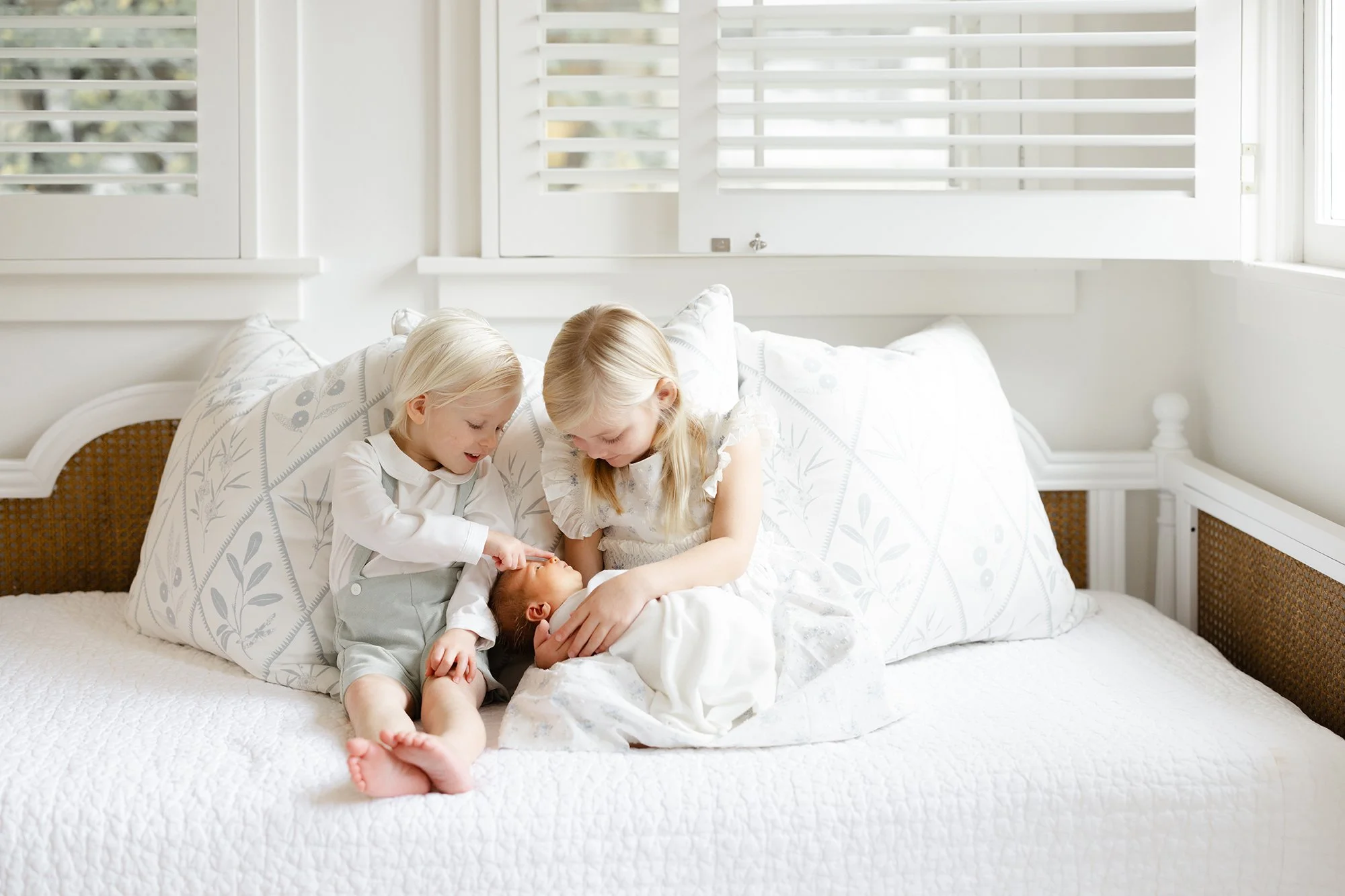 Two young siblings sitting together on a bed beside their newborn baby during a lifestyle newborn session.