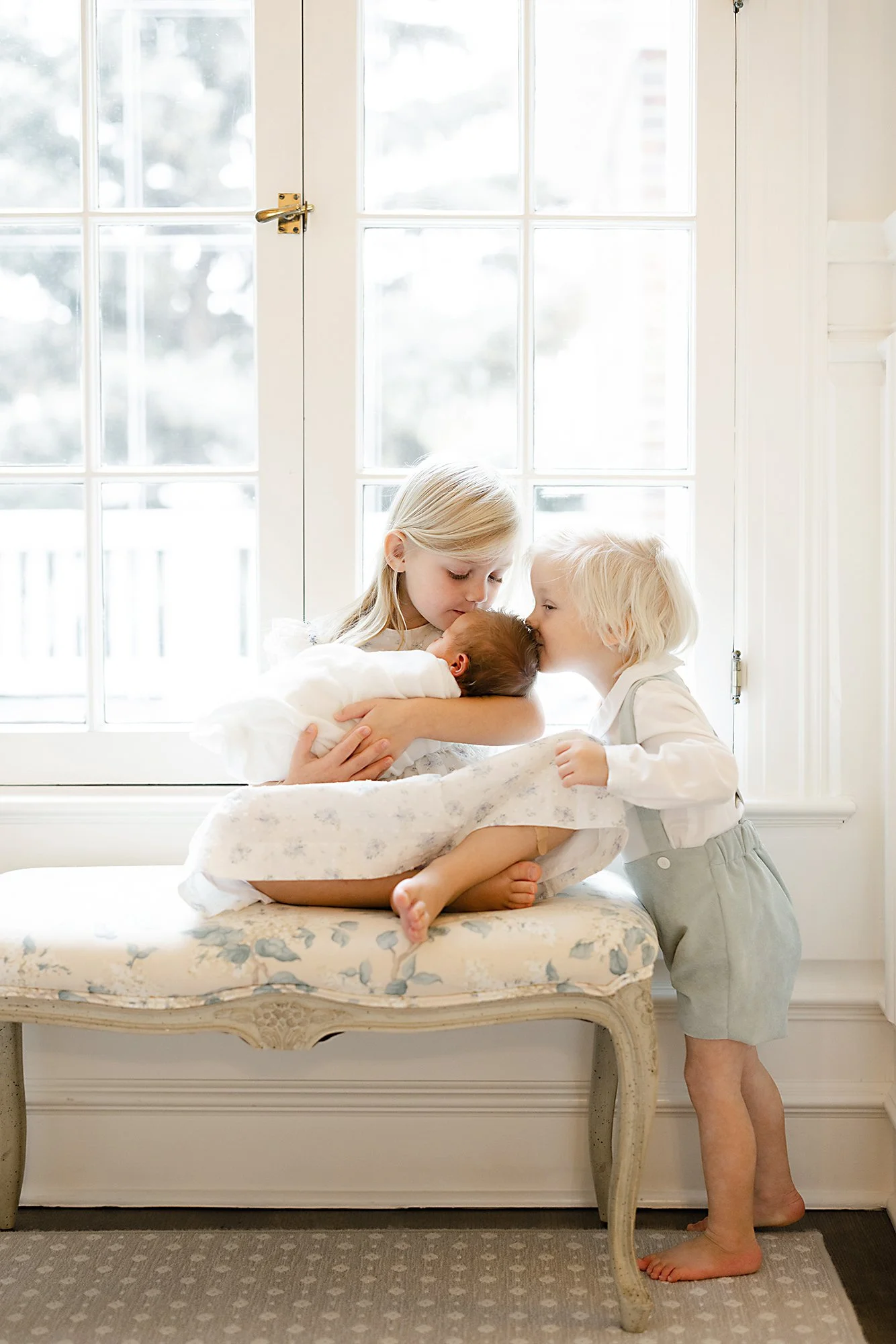 Older siblings gathered around a newborn on a window bench filled with soft natural light.