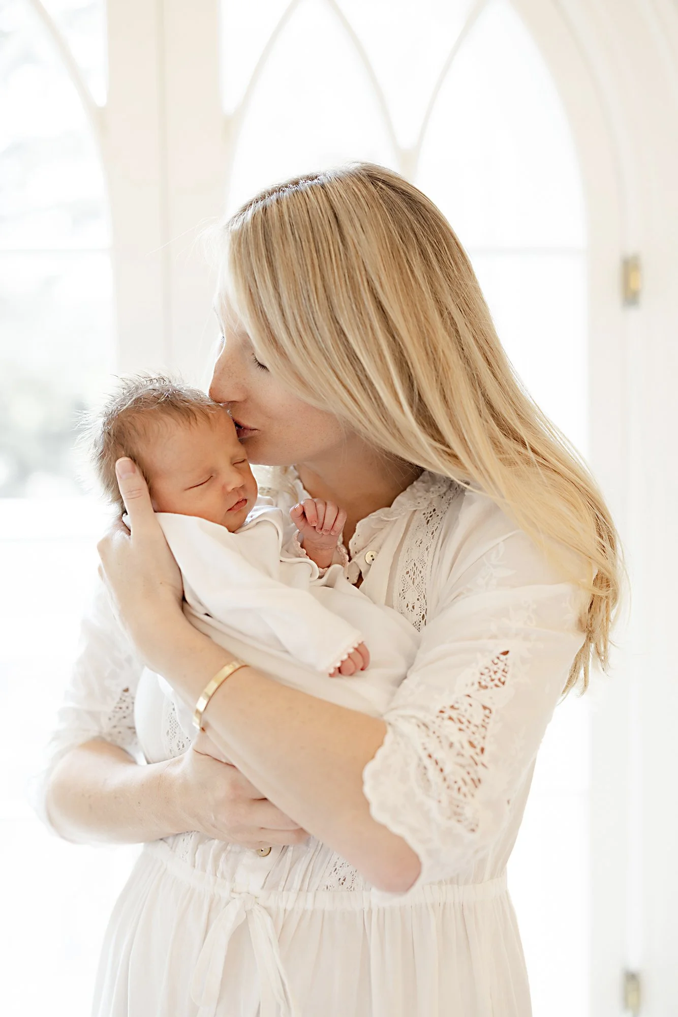 Mother gently kissing her sleeping newborn while standing in soft window light during an in-home newborn portrait session.