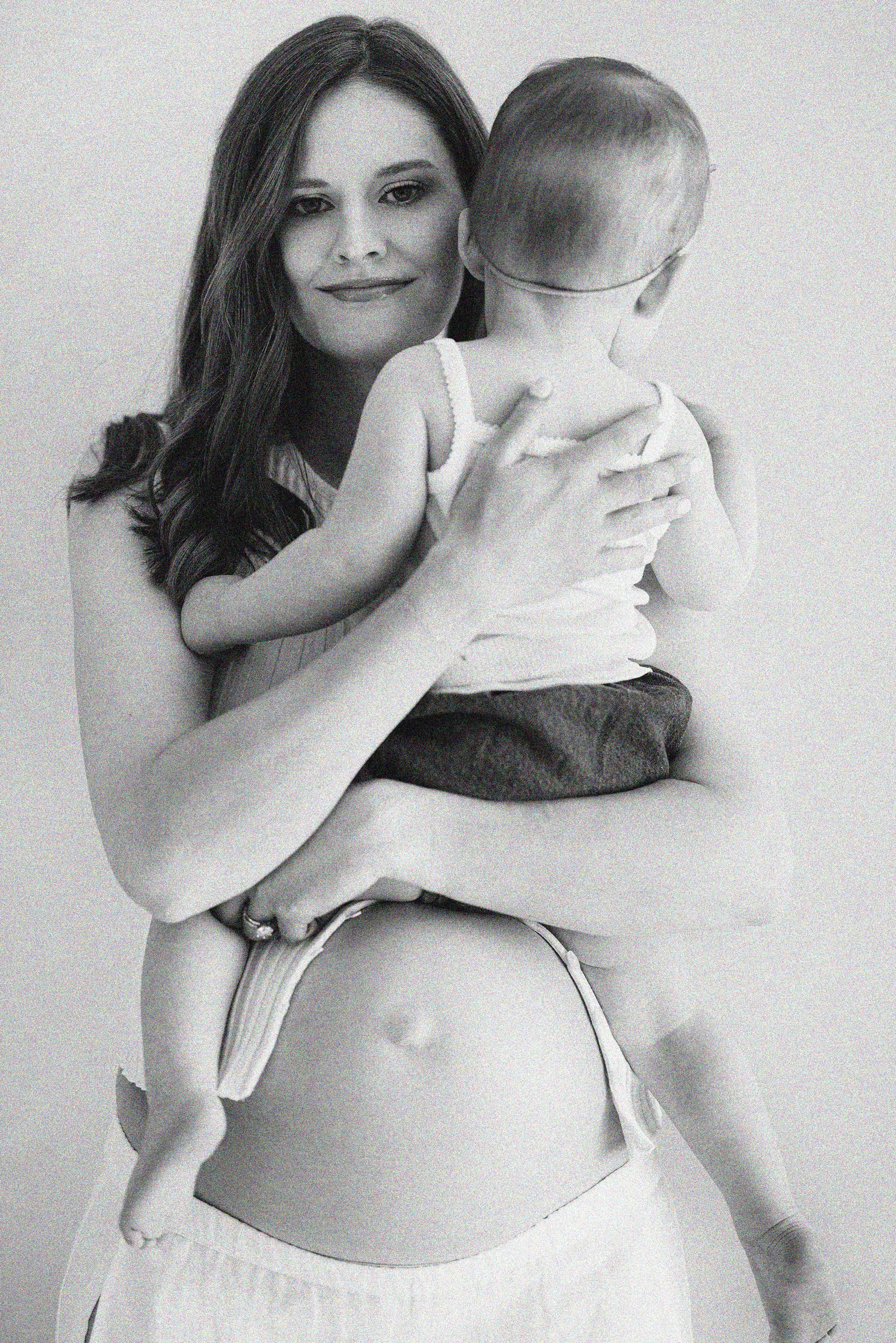 Pregnant mother holding her toddler during a black and white studio maternity portrait.