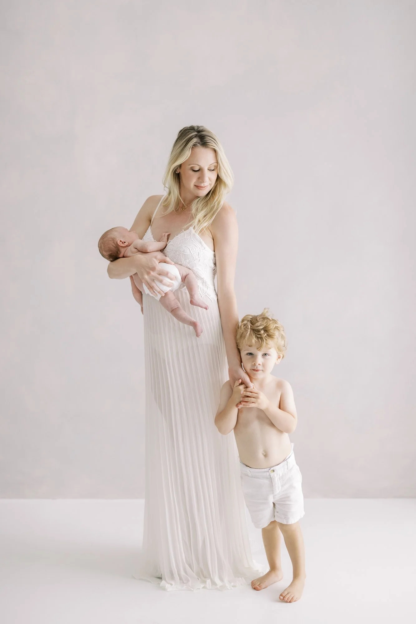 Mother standing with her newborn while her toddler stands beside her during a minimalist family portrait.
