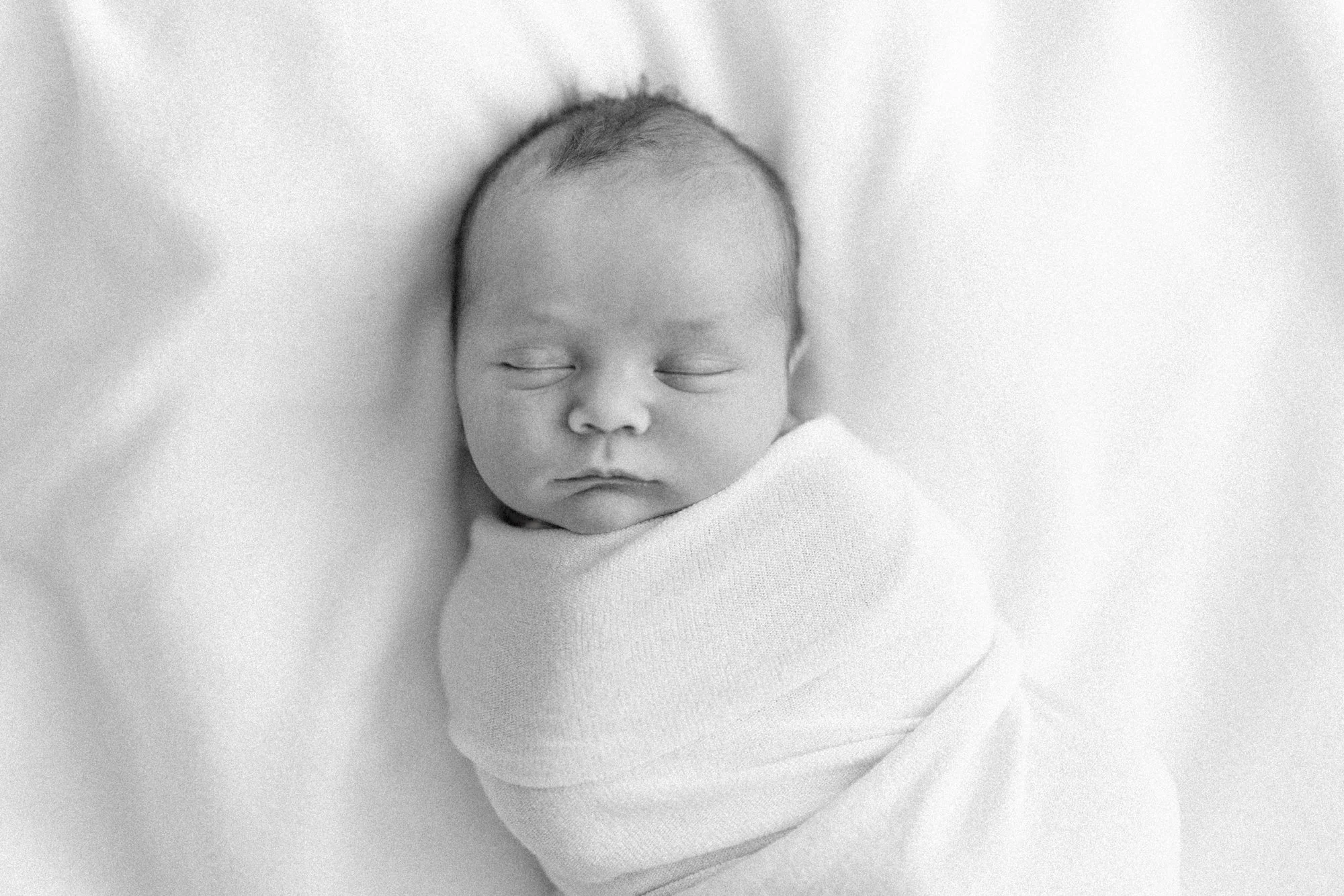Overhead portrait of a sleeping swaddled newborn resting on a soft white blanket during a studio newborn session.