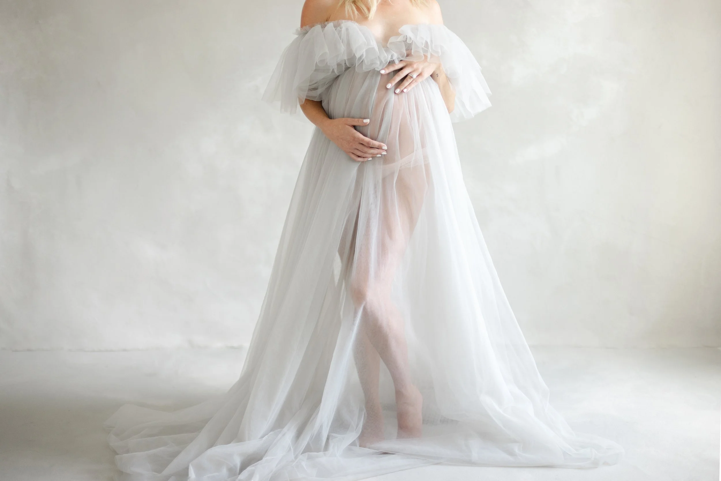 Fine art maternity portrait of an expectant mother wrapped in a flowing white tulle gown with her hands gently resting on her baby bump.