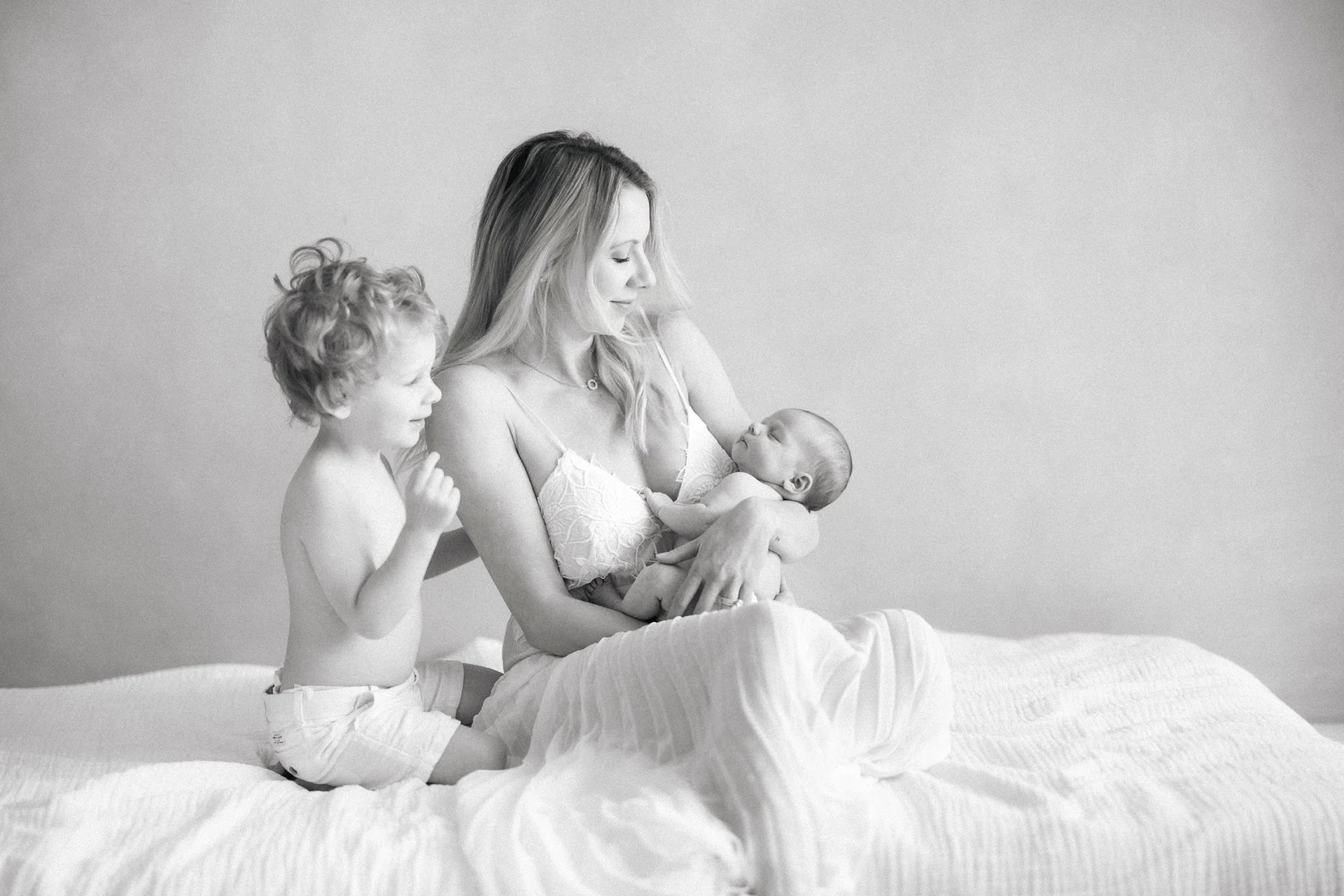 Mother sitting on a bed with her newborn while her toddler sits nearby during a relaxed family newborn session.