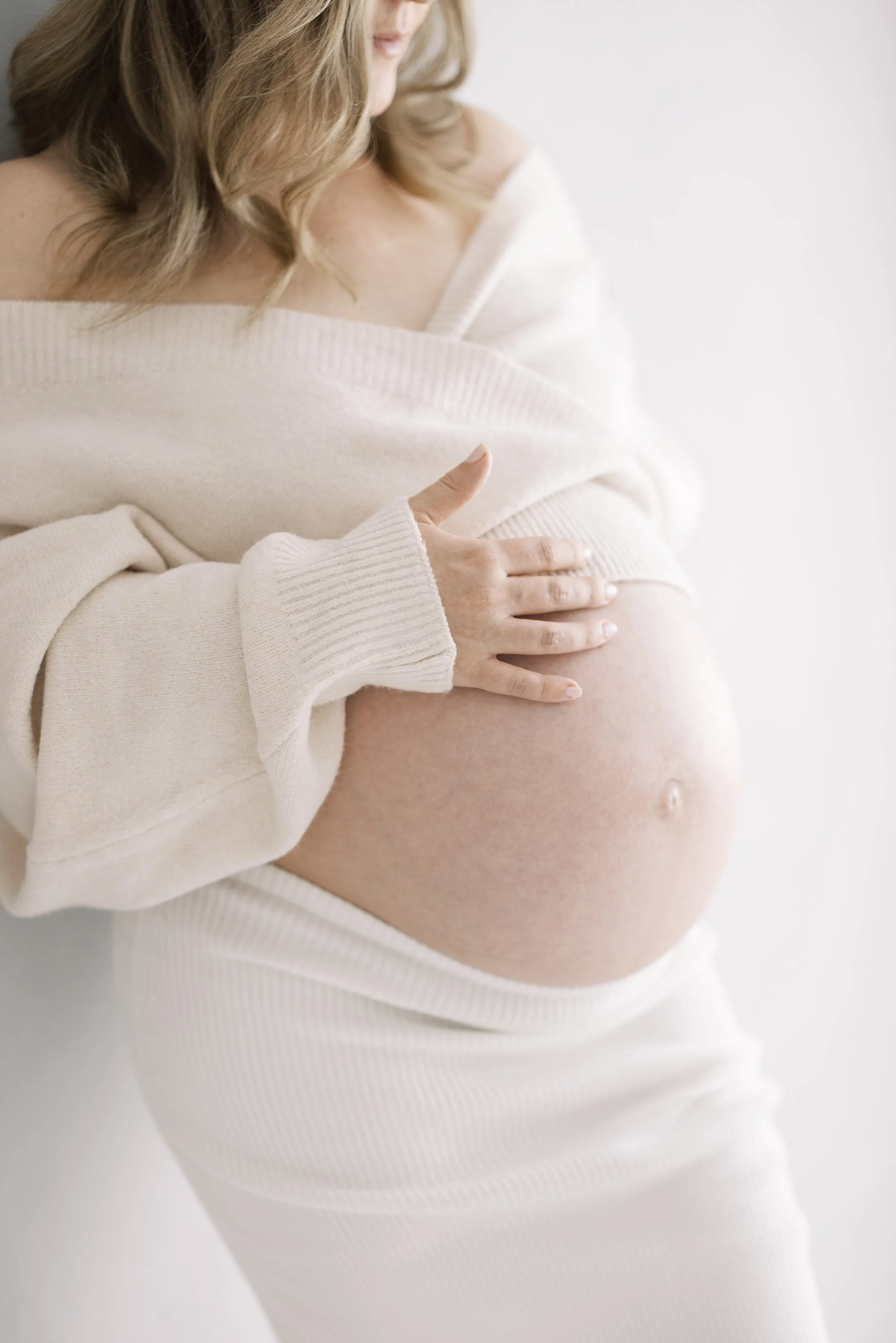 Close-up of an expectant mother wearing a cream knit sweater with her hand gently resting on her baby bump.
