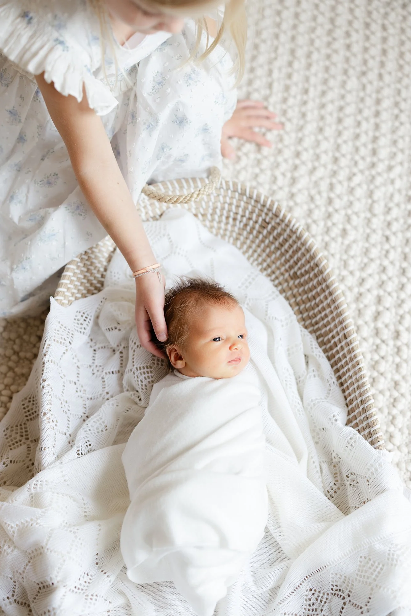 Overhead portrait of a swaddled newborn lying on a bed while an older sister gently reaches toward the baby.