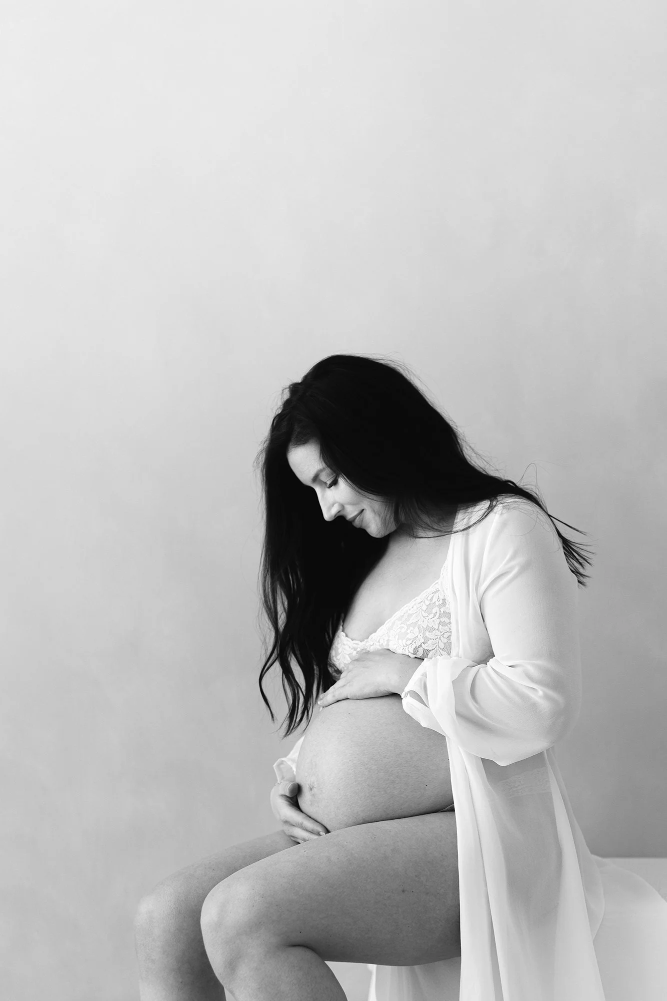 Black and white maternity portrait of an expectant mother looking down at her baby bump while wearing a lace bralette and sheer robe.