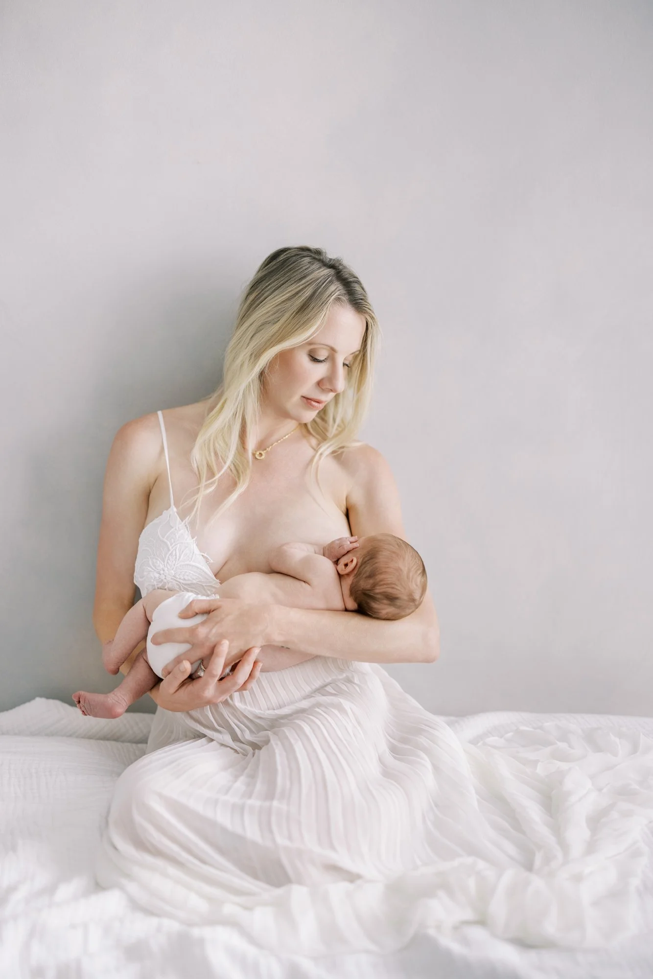 Mother seated on a bed while gently holding her newborn during a studio newborn photography session.
