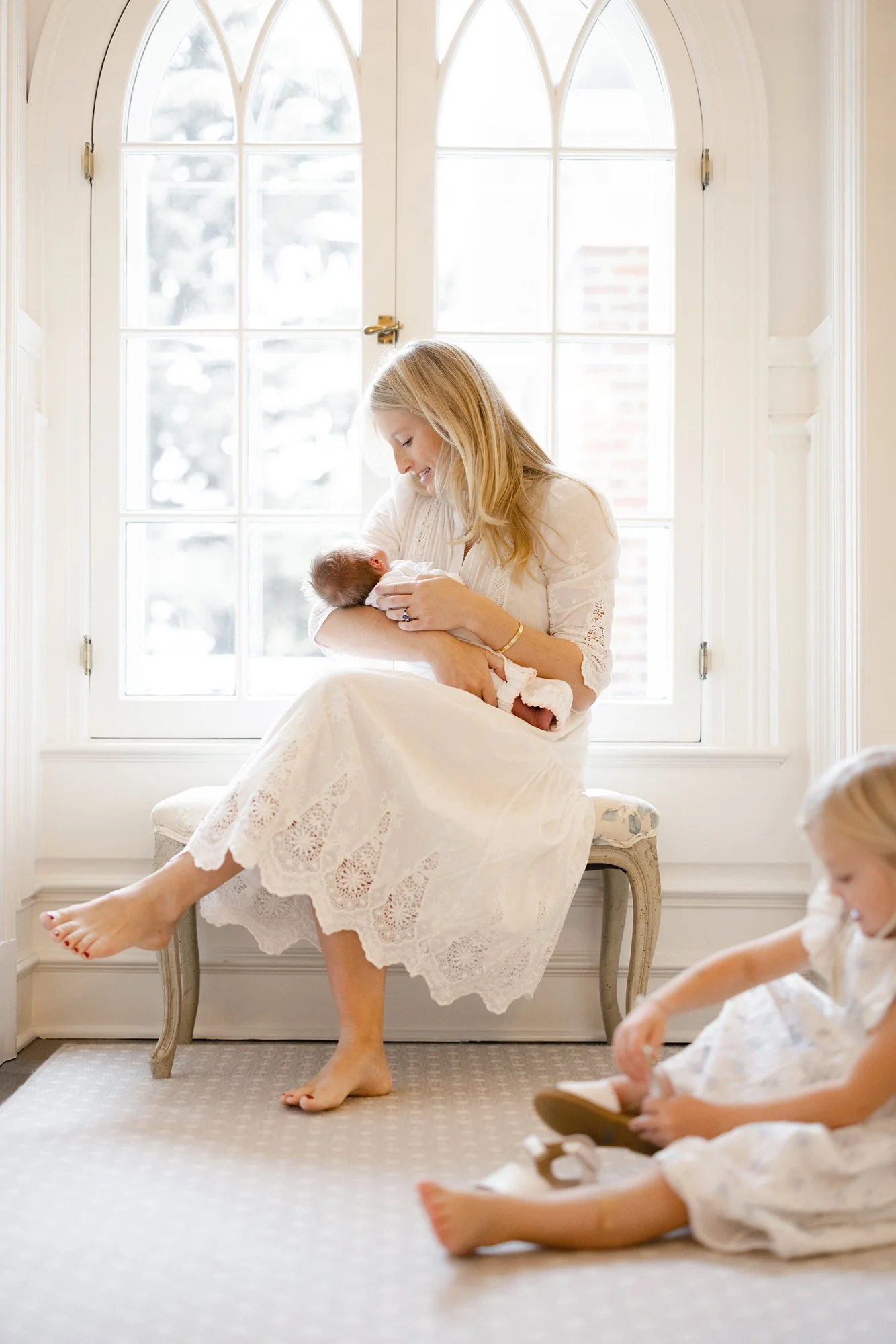 Mother holding her newborn beside a large window while older children play nearby.