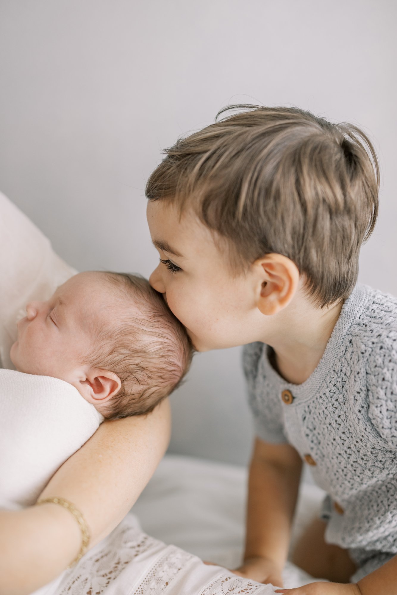 Older brother gently kissing his newborn sibling during a family newborn photography session.
