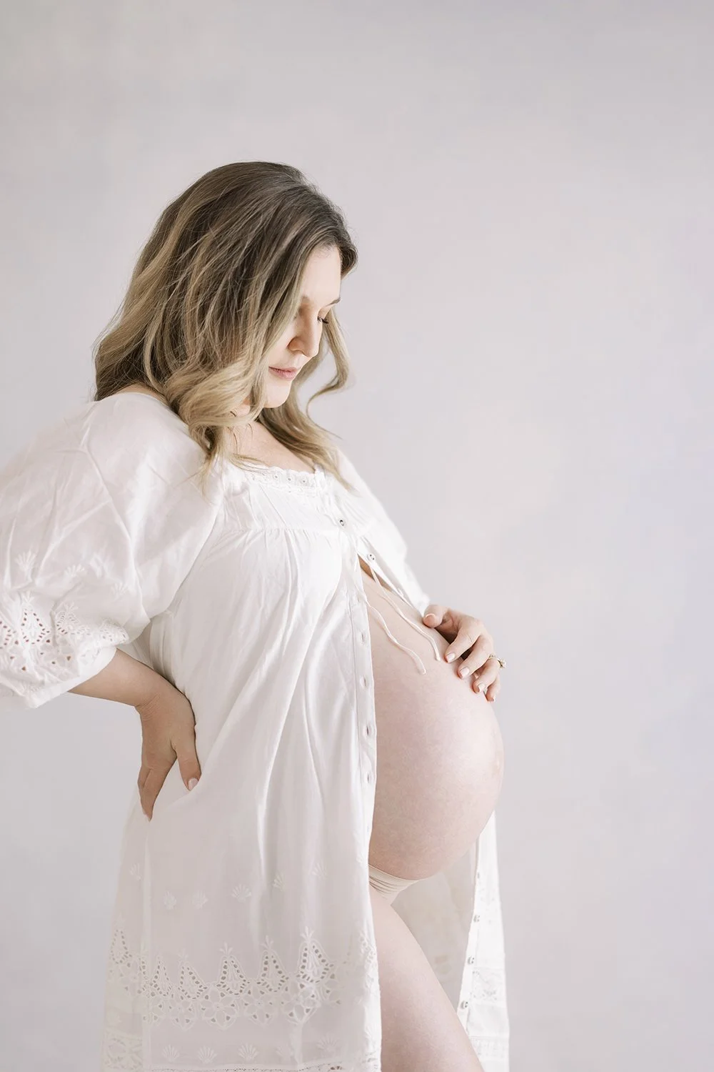 Pregnant mother wearing a white lace robe looking down at her baby bump during a studio maternity session.