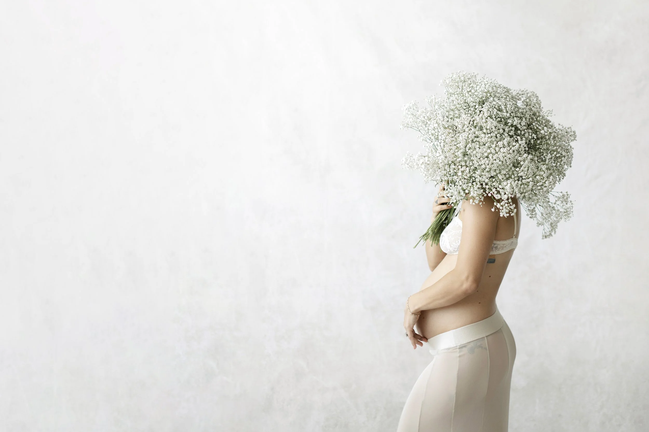 Pregnant woman holding baby’s breath flowers in front of her face during an artful studio maternity portrait session.