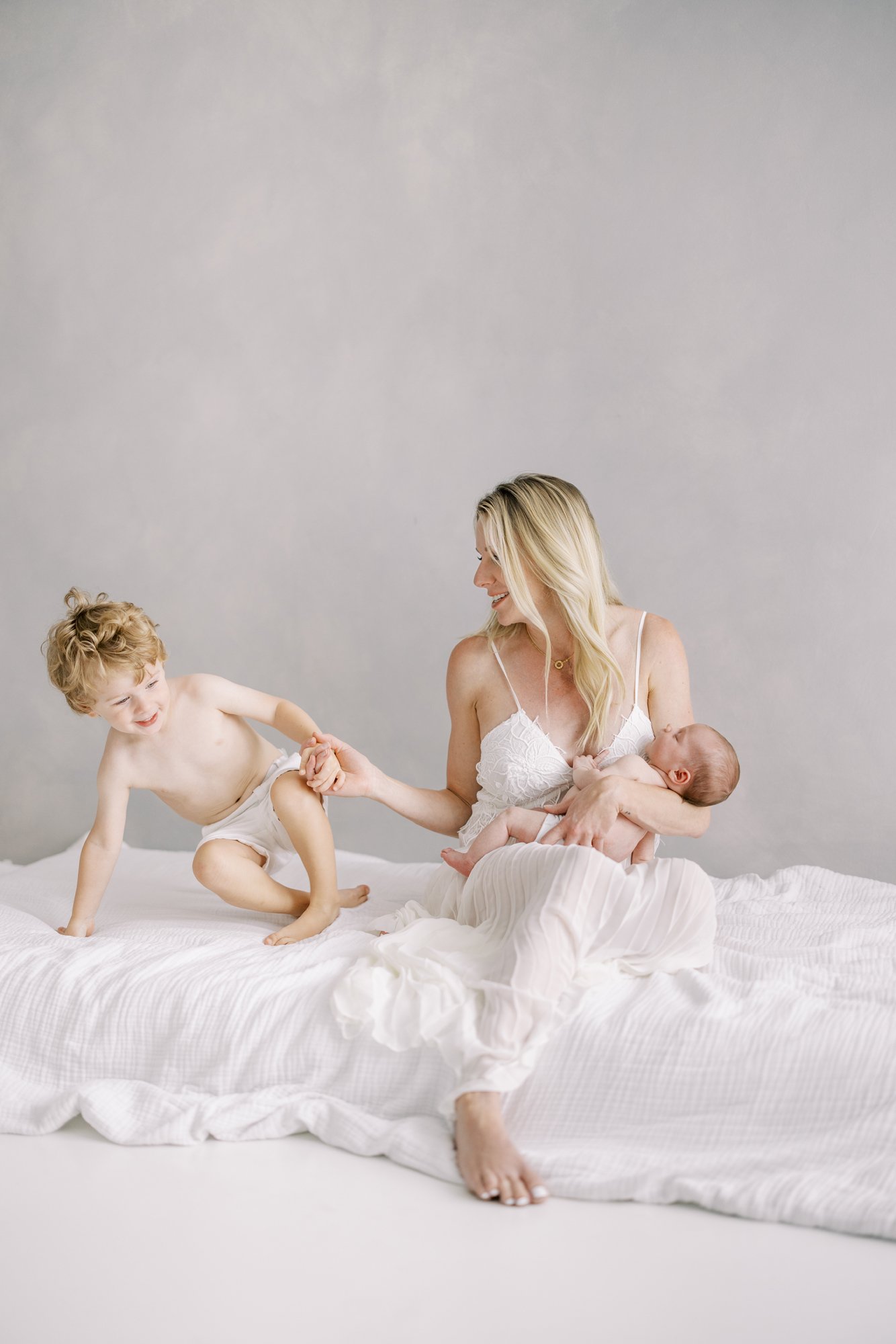 Mother sitting with her newborn while interacting with her toddler on a bed during a relaxed family session.