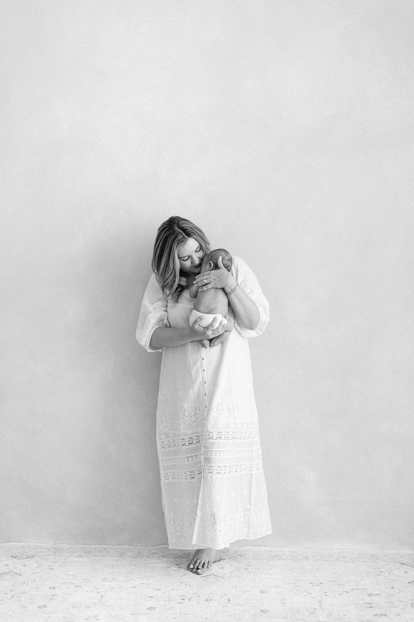Black and white portrait of a mother standing while holding her newborn in a minimalist studio.