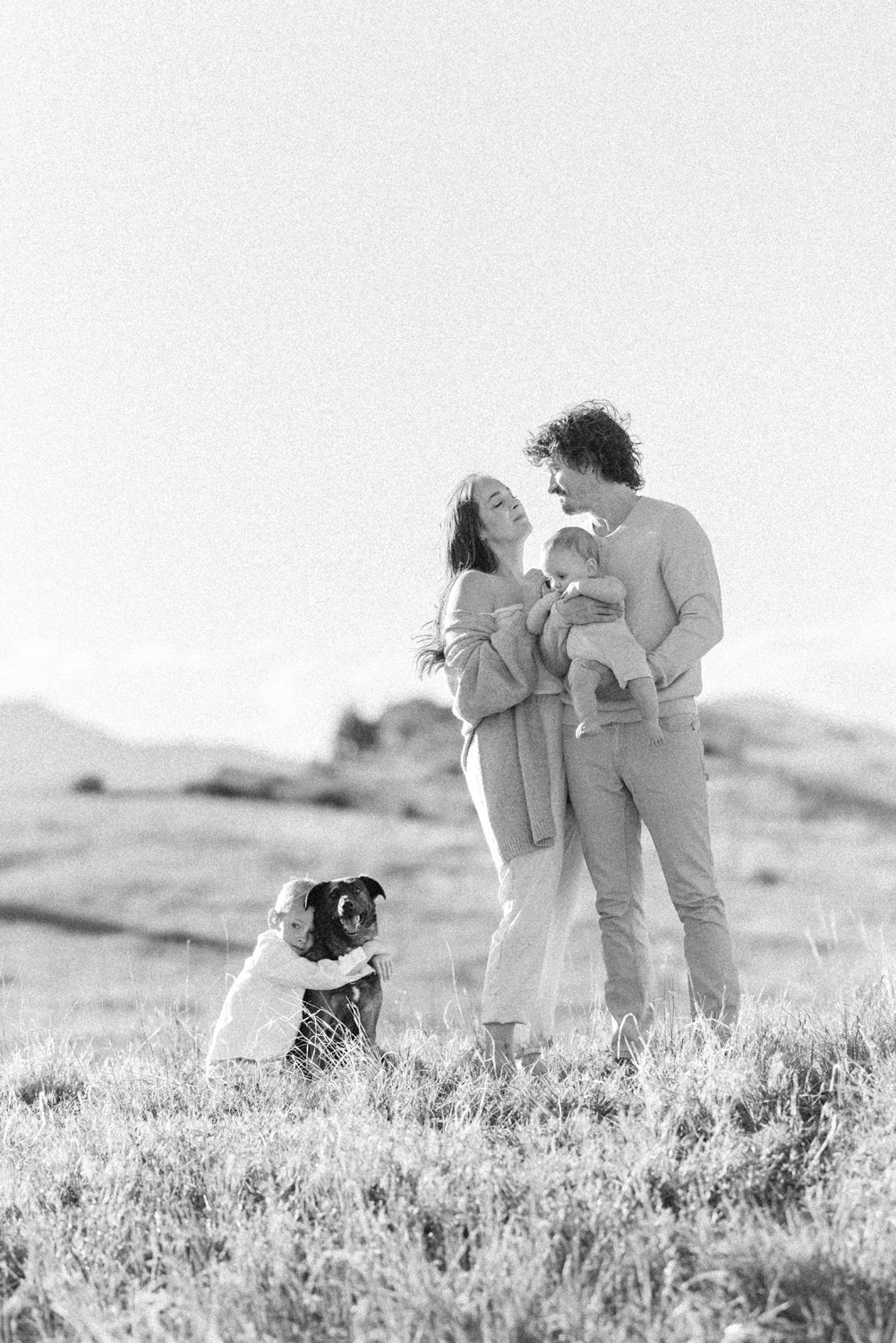 Black-and-white portrait of parents embracing with their children playing around them in a Colorado field.
