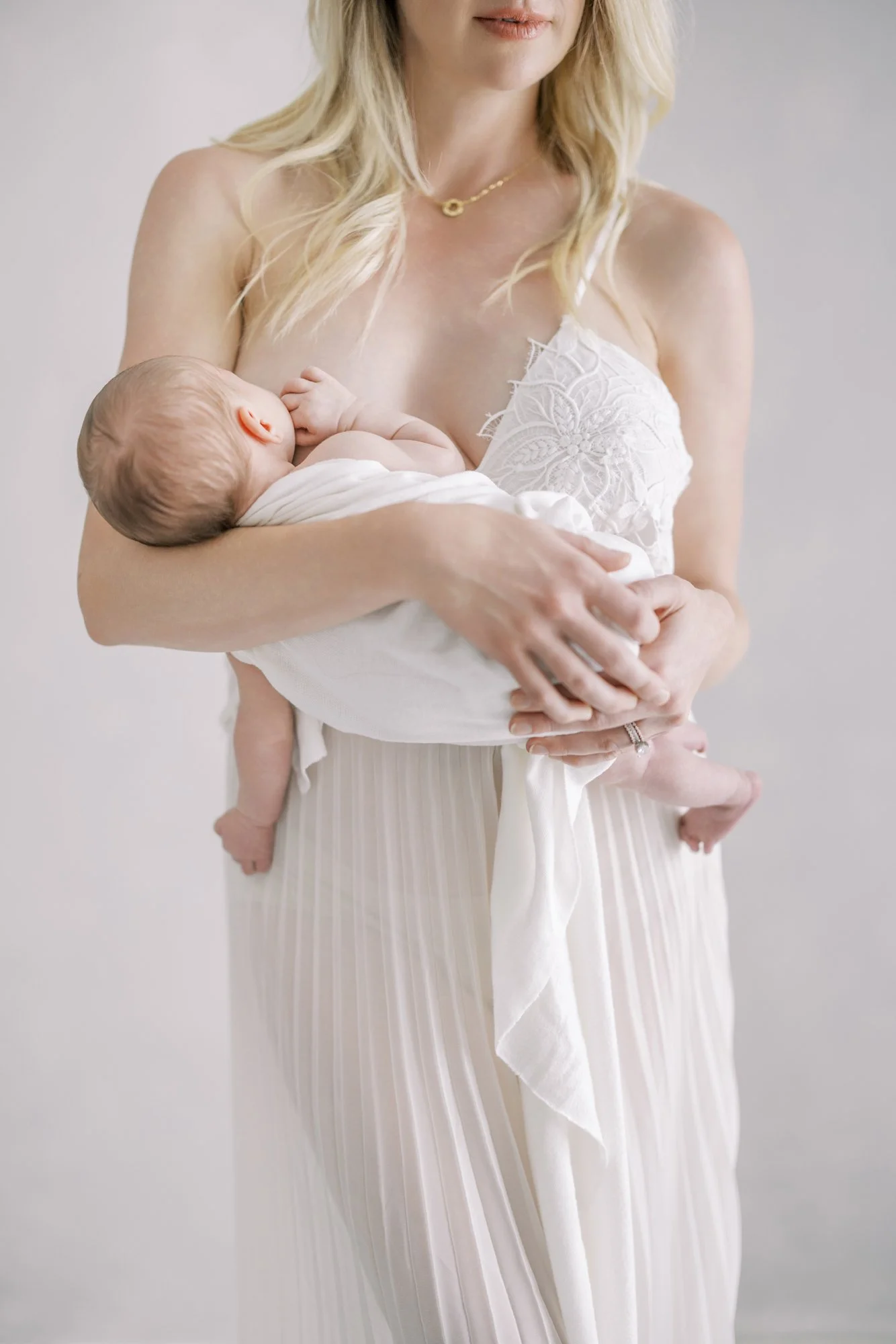 Mother seated on a bed while gently holding her newborn during a studio newborn photography session.