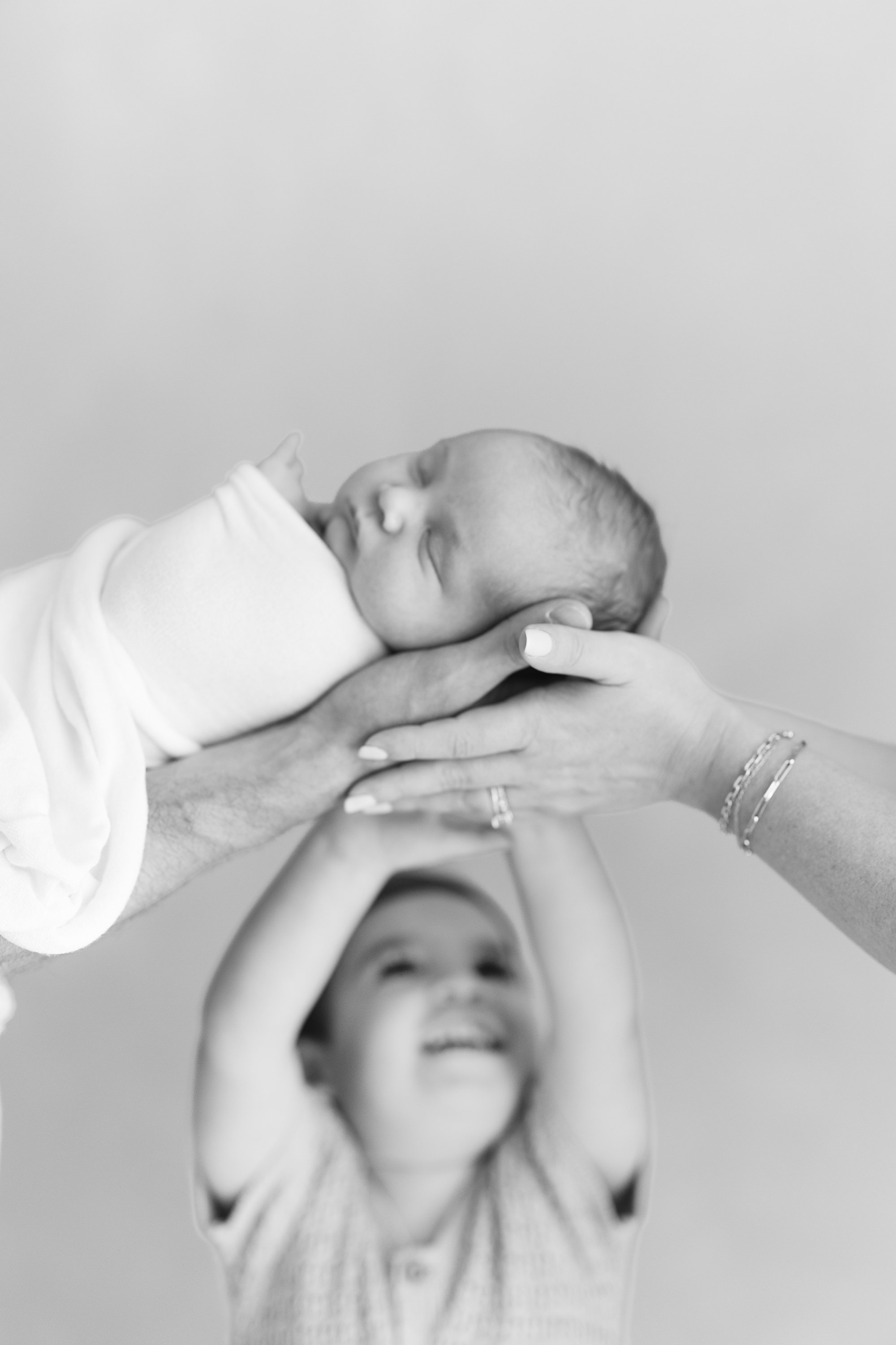 Older sibling reaching up toward a newborn being gently held overhead during a playful family portrait.