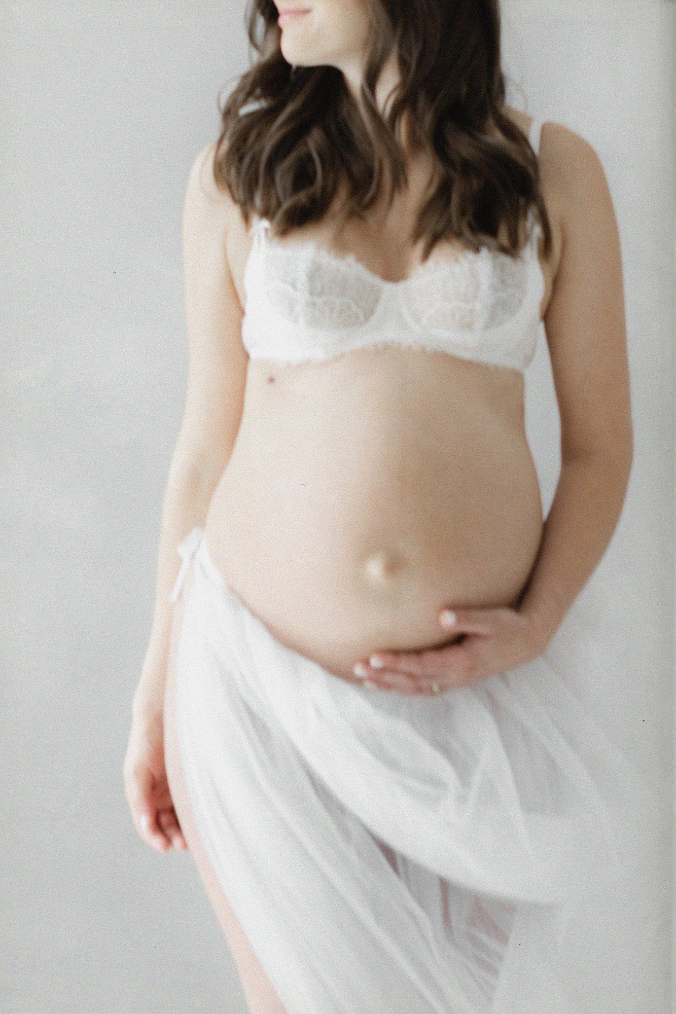 Close-up maternity portrait of an expectant mother wearing a white lace bralette and flowing tulle skirt with her hand resting on her baby bump.