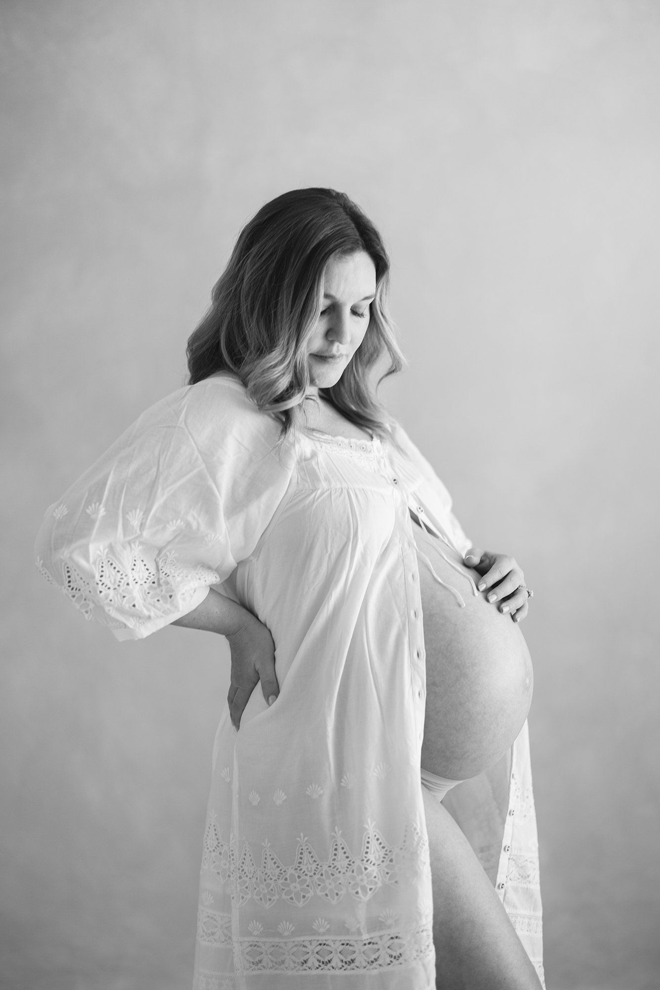 Pregnant mother in a white lace robe looking down at her belly during a studio maternity session in Denver.