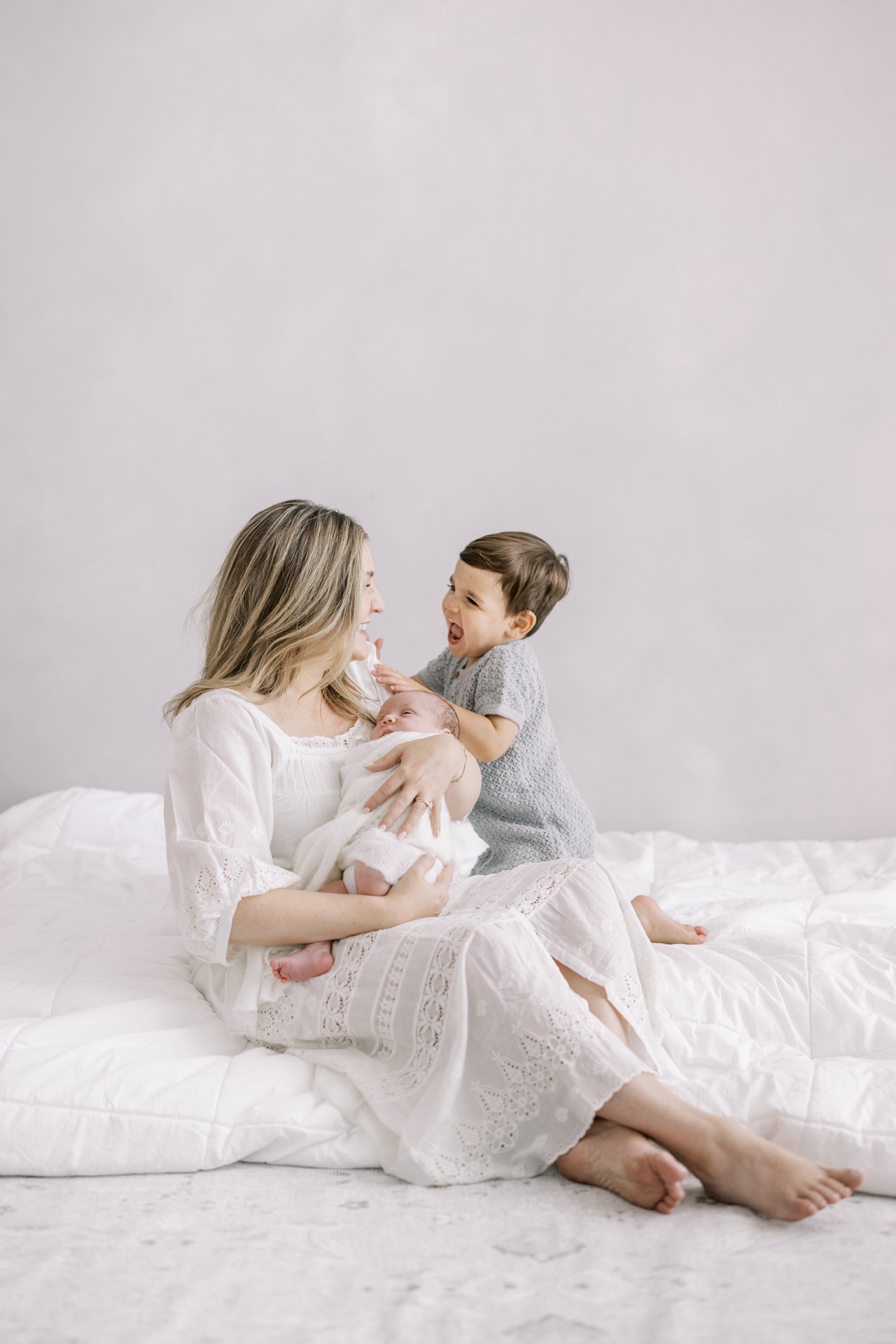 Mother sitting on a bed with her toddler while holding their newborn during a relaxed family newborn session.