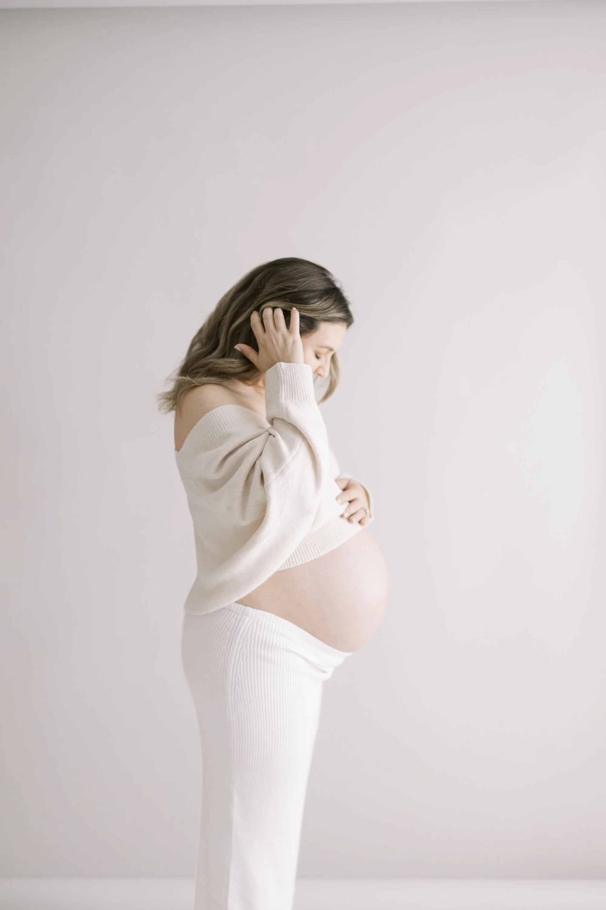 Expectant mother standing in profile wearing a cream knit sweater and long white skirt during a minimalist studio maternity session.