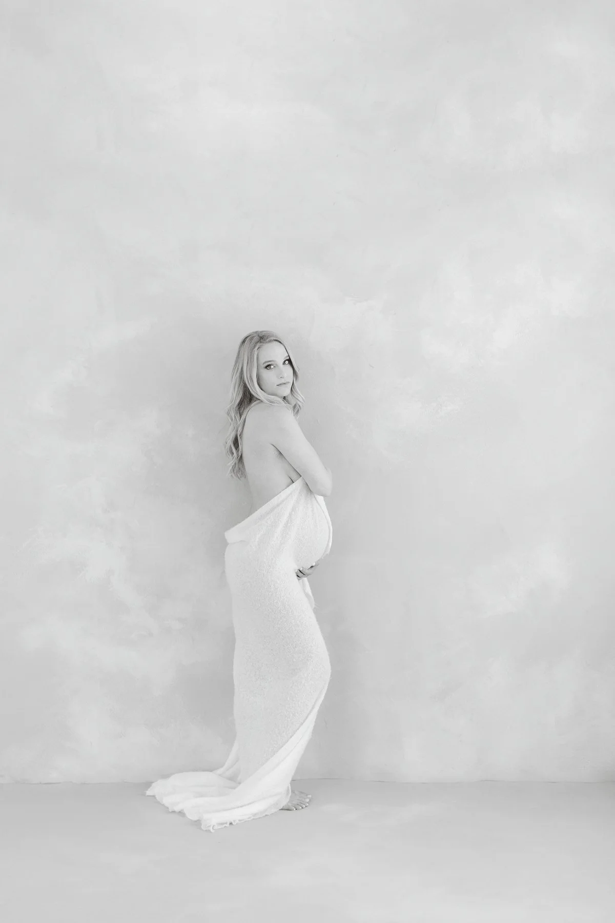 Black and white full-length portrait of a pregnant mother wrapped in a flowing white drape while looking over her shoulder.