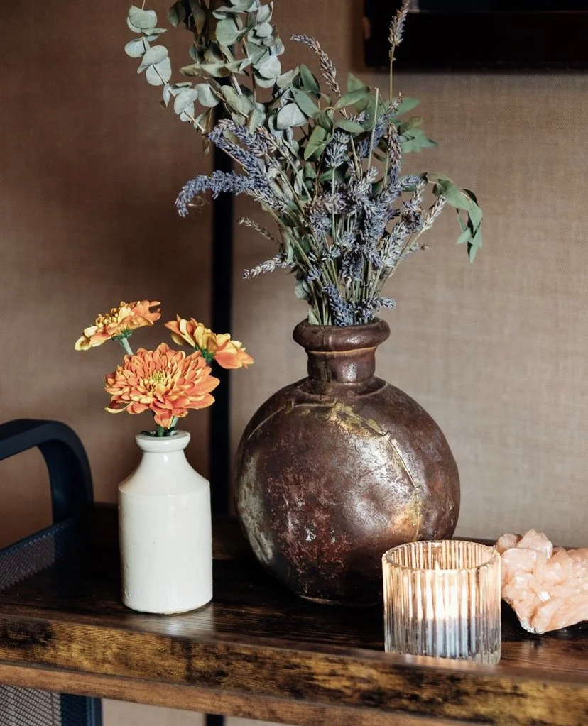 A rustic scene with two vases and a lit candle on a wooden surface. One vase is round and brown, filled with dried lavender and eucalyptus. The other vase is white with orange flowers. A candle in a glass holder is lit, and pink crystals are placed nearby.
