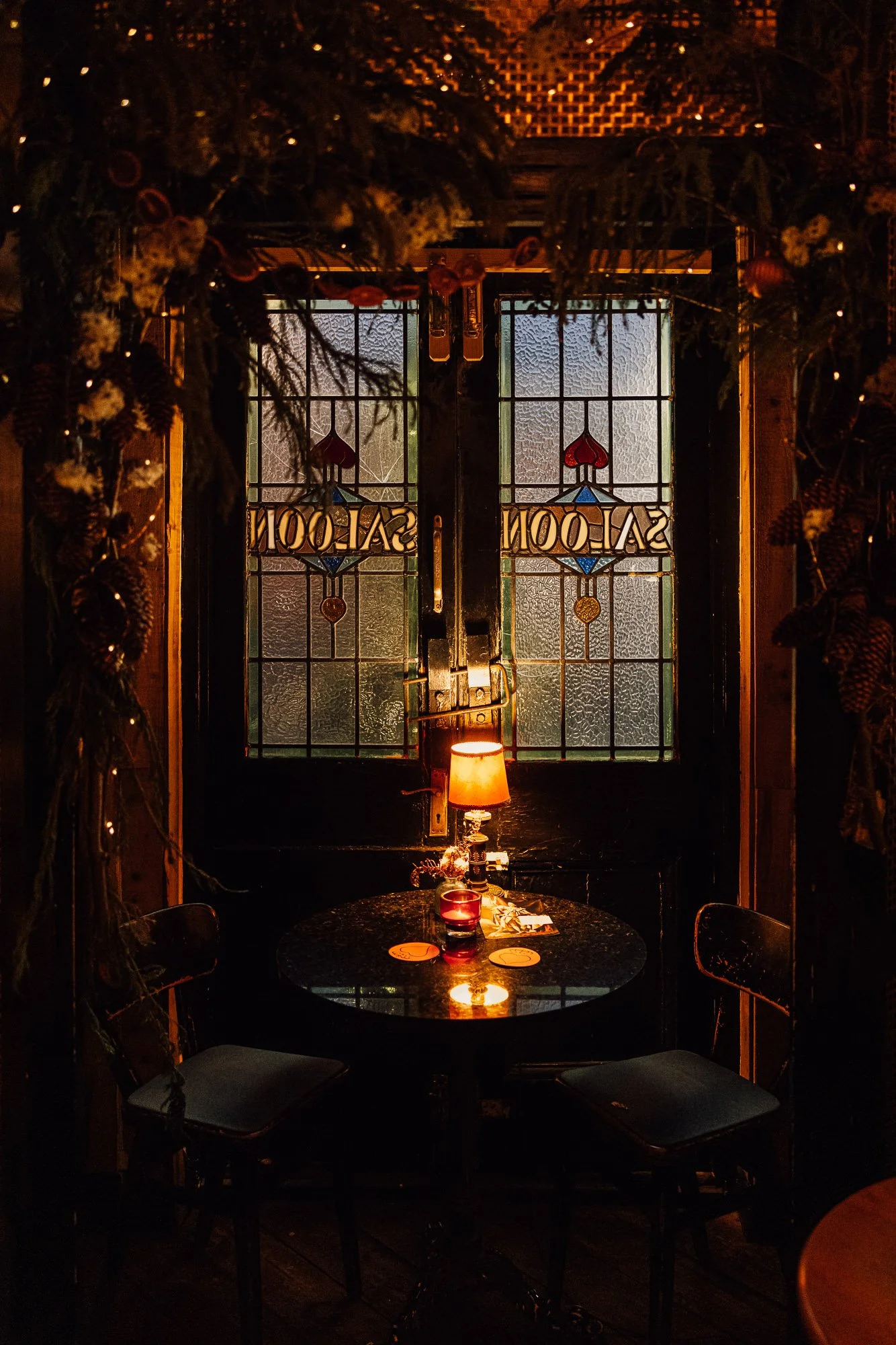 Cozy dimly lit room with stained glass doors labeled 'Saloon,' round table with a small lamp, and two chairs. Decorated with hanging pine cones and greenery.