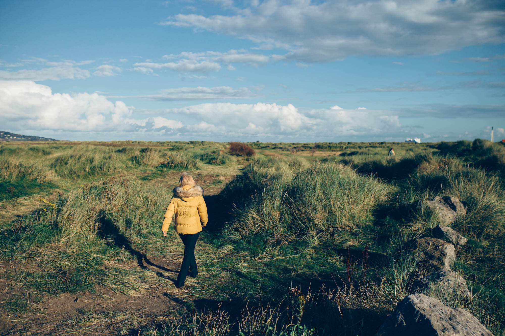 A person wearing a yellow jacket walking along a grassy dirt path through a grassy field, under a partly cloudy sky with patches of blue.