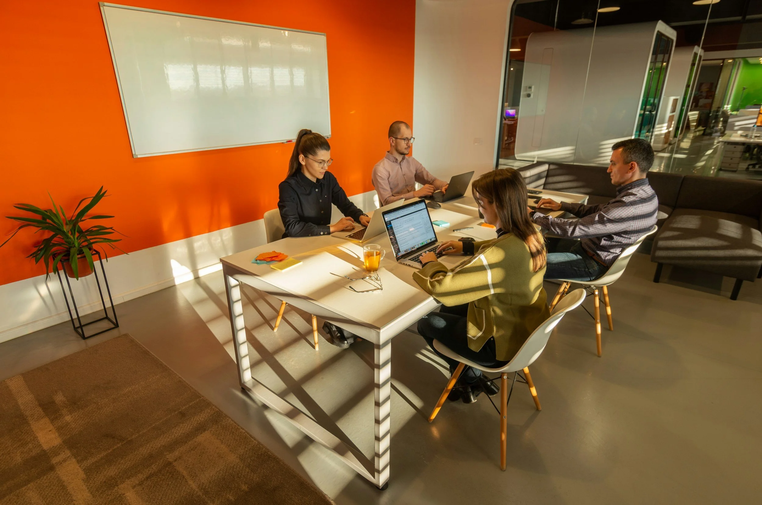 Four coworkers working on laptops at a conference table in a modern office with orange accent wall and large glass partition.