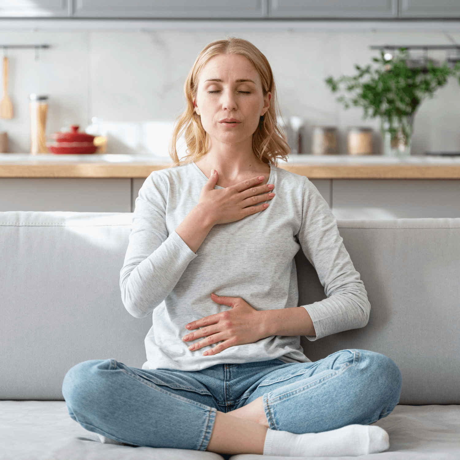 A woman sitting cross-legged on a sofa, with one hand on her chest and the other on her stomach, appearing to breathe deeply with a calm expression.