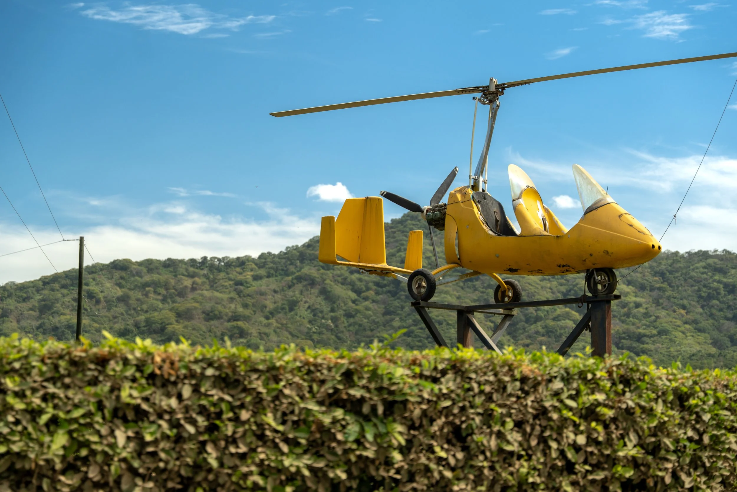 A yellow helicopter mounted on a metal platform outdoors with green hills in the background and utility wires overhead under a blue sky with some clouds.