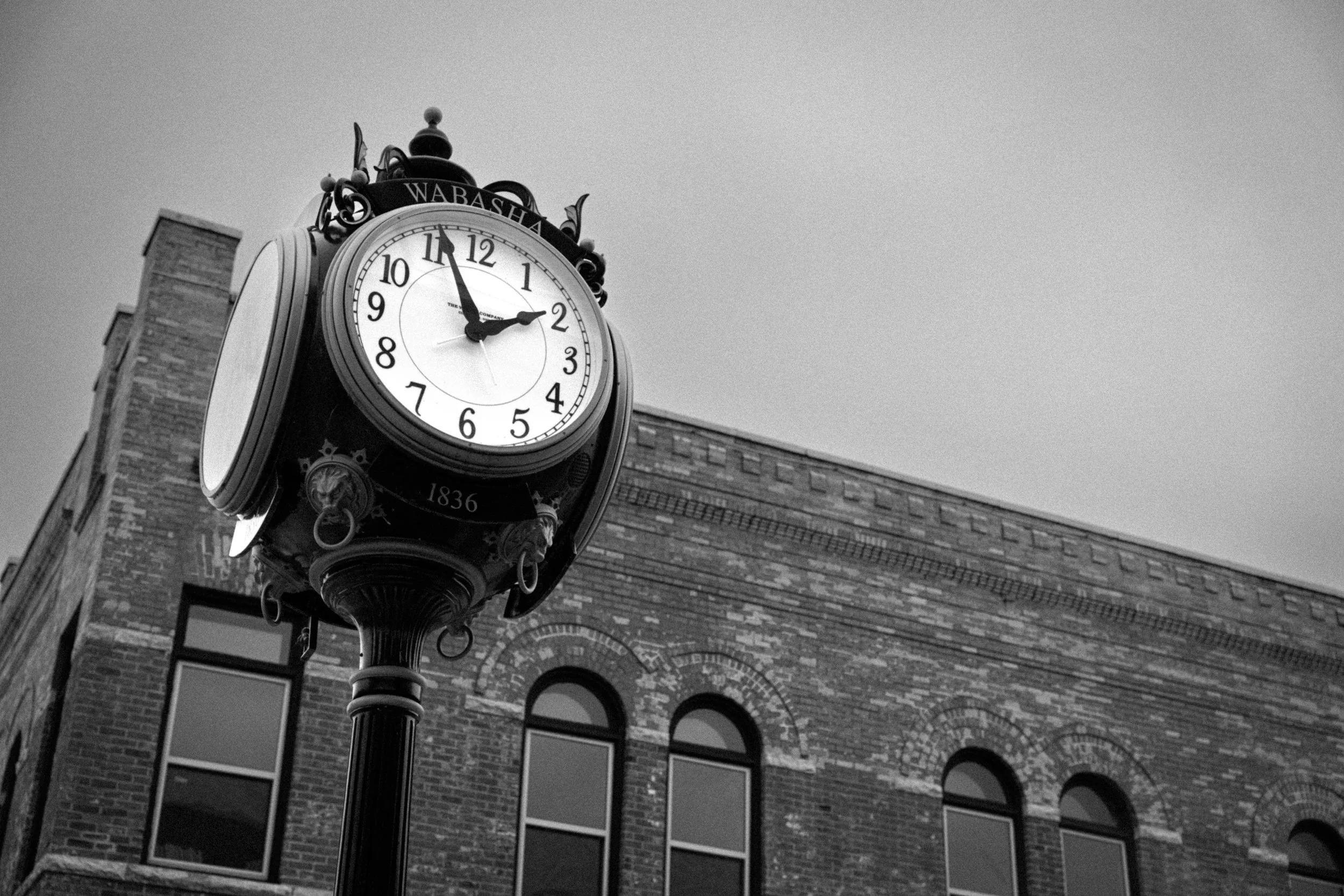 A vintage clock on a tall stand in front of a brick building, with the time showing 2:54.