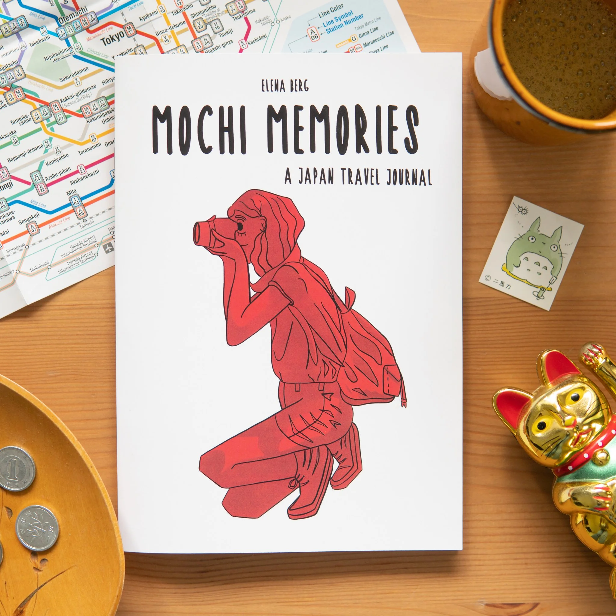 Flatlay of “Mochi Memories” journal surrounded by Japanese travel items: Tokyo subway map, tea, coins, Totoro sticker and Maneki-neko. A warm invitation into personal storytelling and cultural detail.