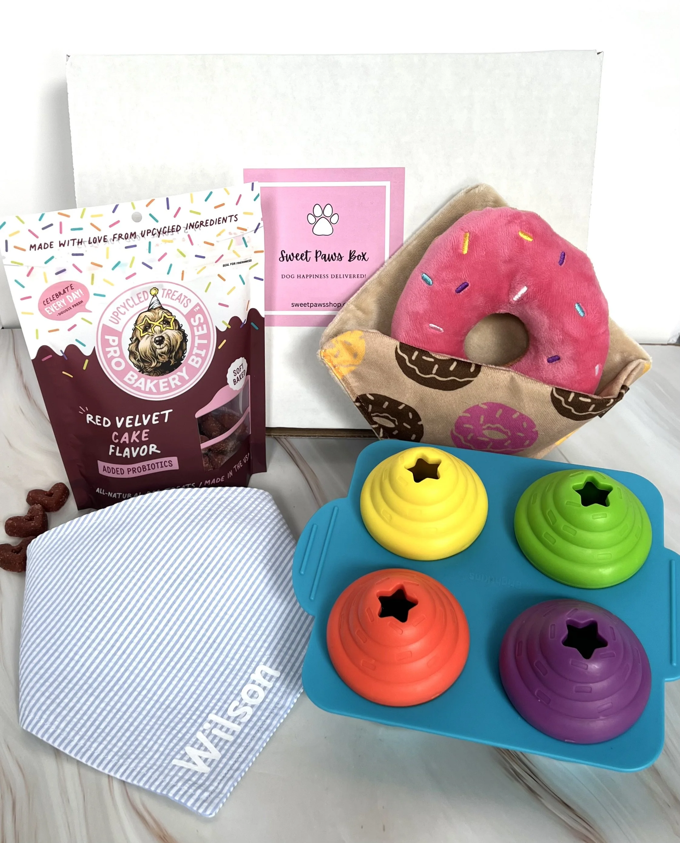 A gift box containing dog treats, a plush donut toy, a colorful puzzle toy with four rotating sections, a branded cloth, and a pink card from Sweet Paws Box.