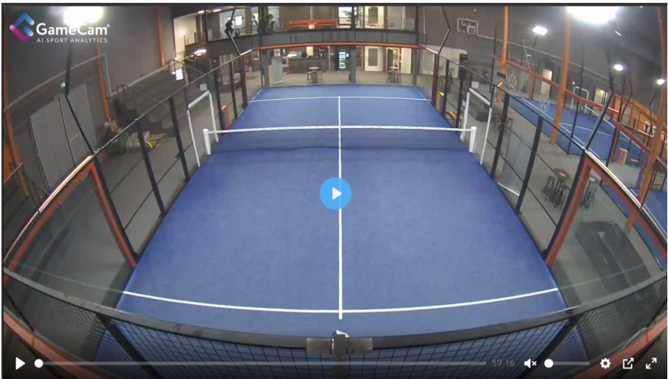 Indoor padel court with blue surface and glass walls, illuminated lighting, and sitting areas outside the court.