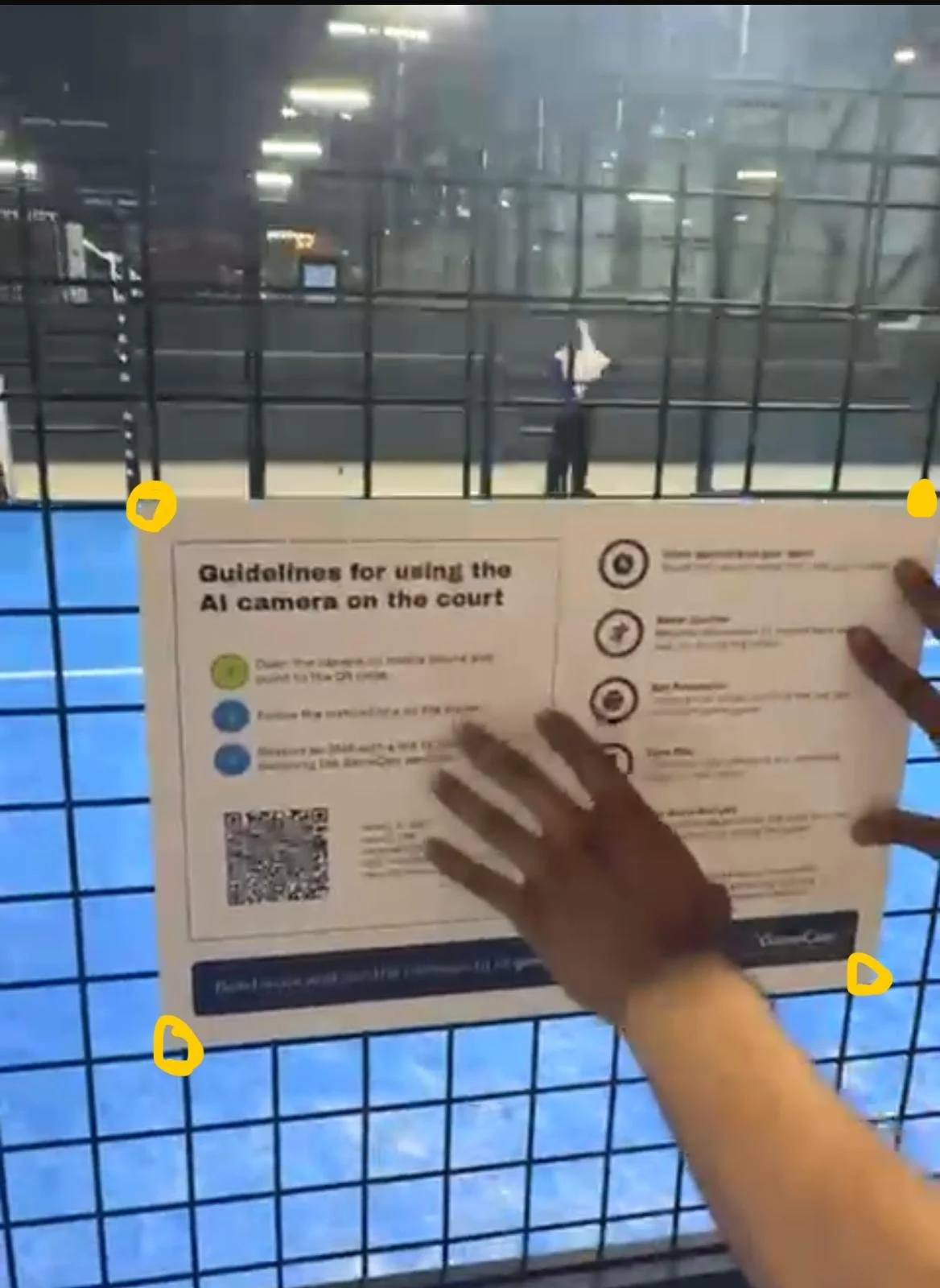 Sign with guidelines for using AI camera on a sports court, displayed on a wire fence at an indoor court with a person in the background.