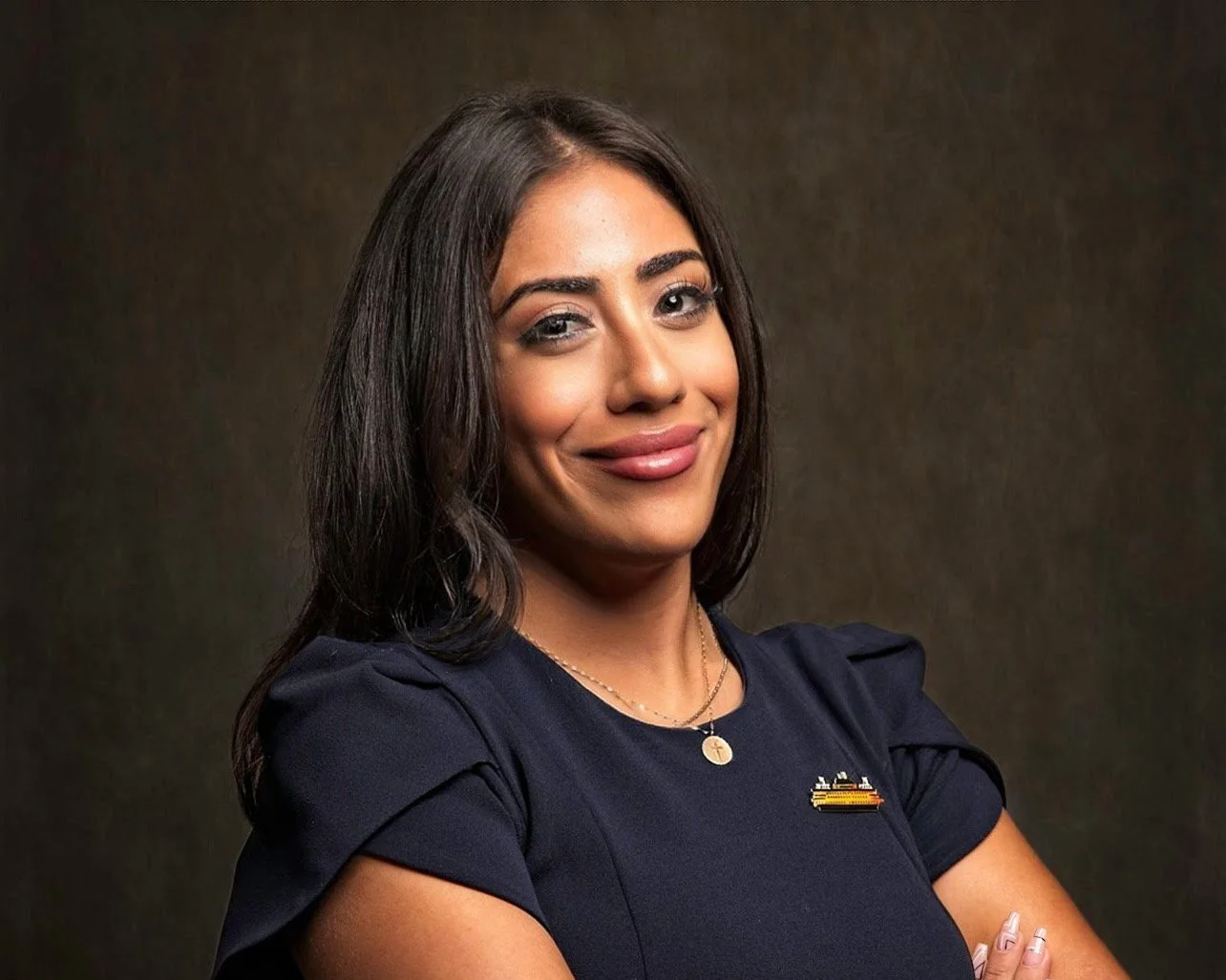 Portrait of a woman with long dark hair, wearing a navy blue top with a small flag pin, gold necklace, and smiling with crossed arms against a dark background.
