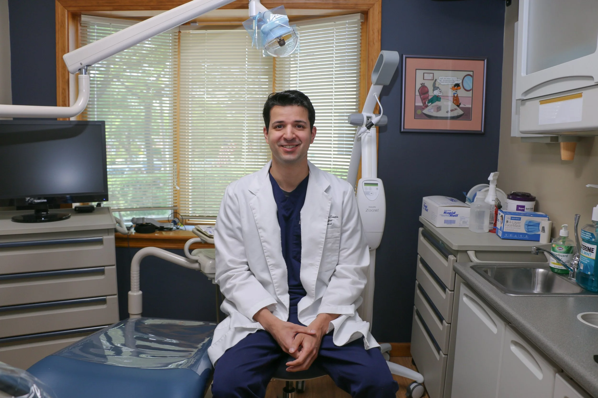 A smiling male healthcare professional sitting in a medical examination room, surrounded by medical equipment, with a window and blinds behind him.