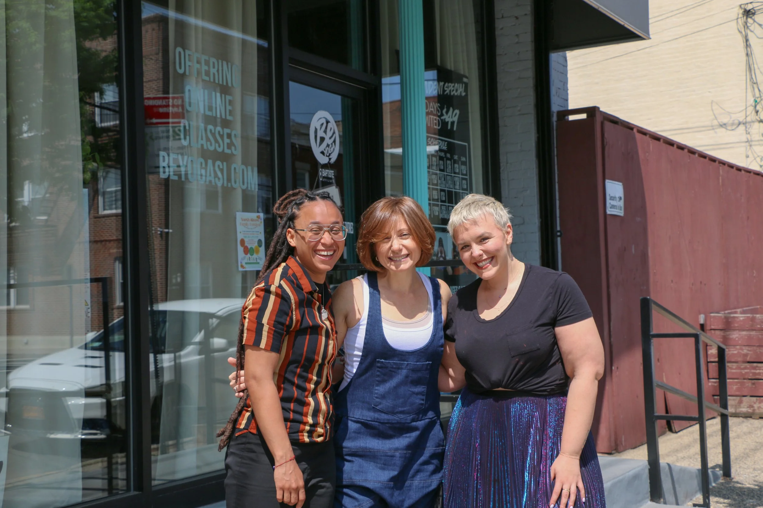 Three women standing outside a building, smiling at the camera, with large glass windows and a sign that reads offering online classes at beygasi.com.