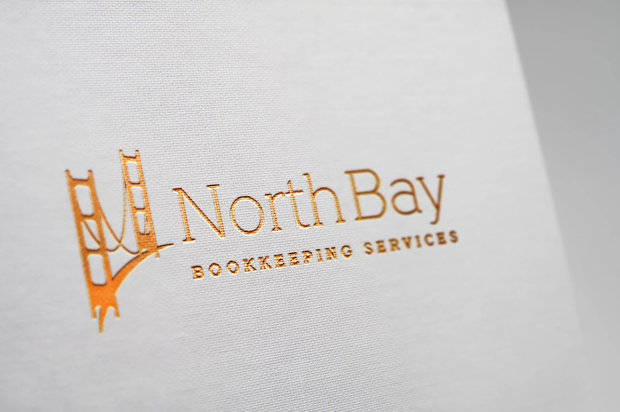 Close-up of a business card or brochure with a gold foil logo and text reading 'North Bay Bookkeeping Services' and an illustration of a bridge.