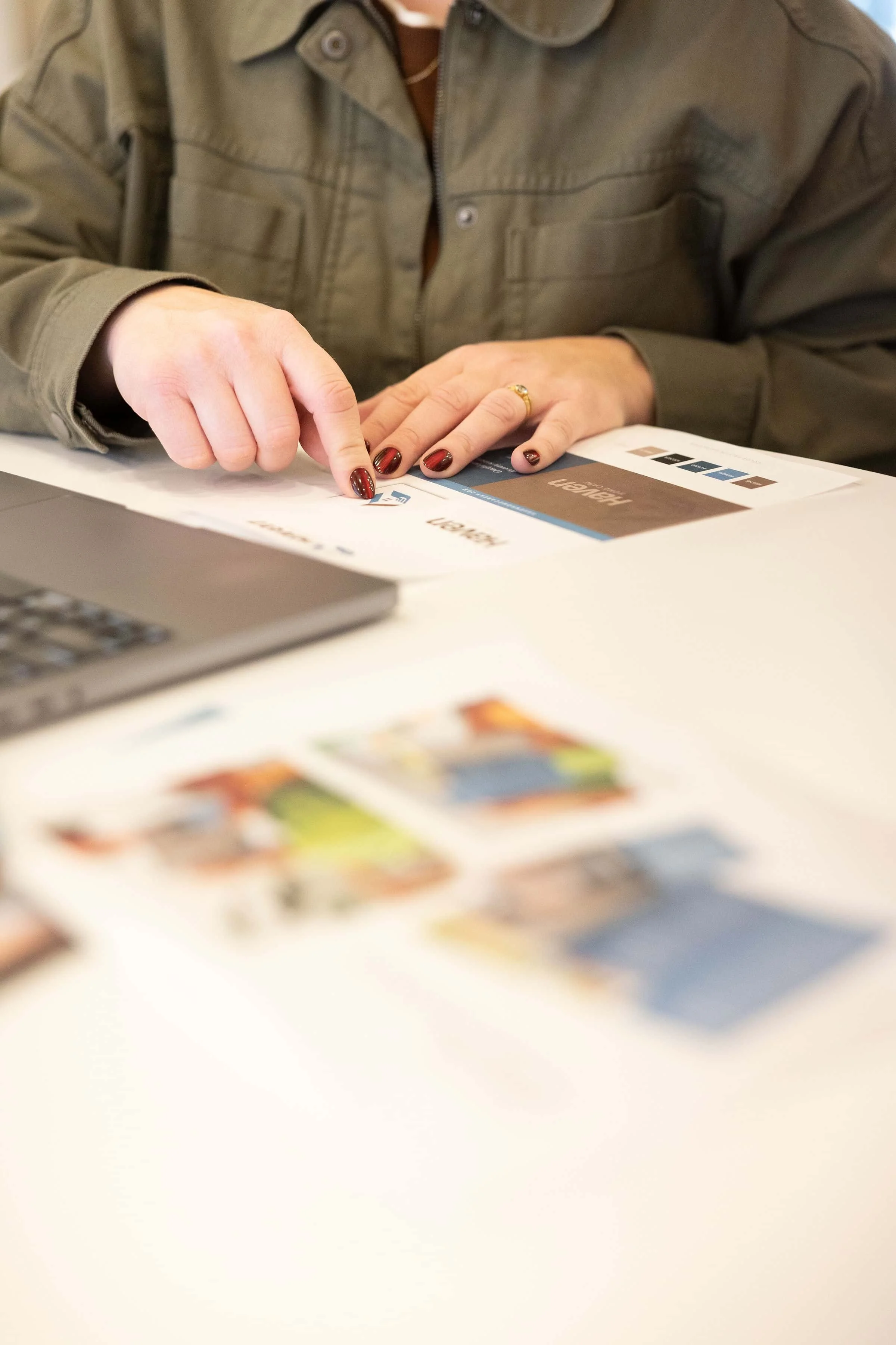 A person with painted nails reviews a color palette and design catalog on a white table, with a laptop and colorful brochures in the foreground.