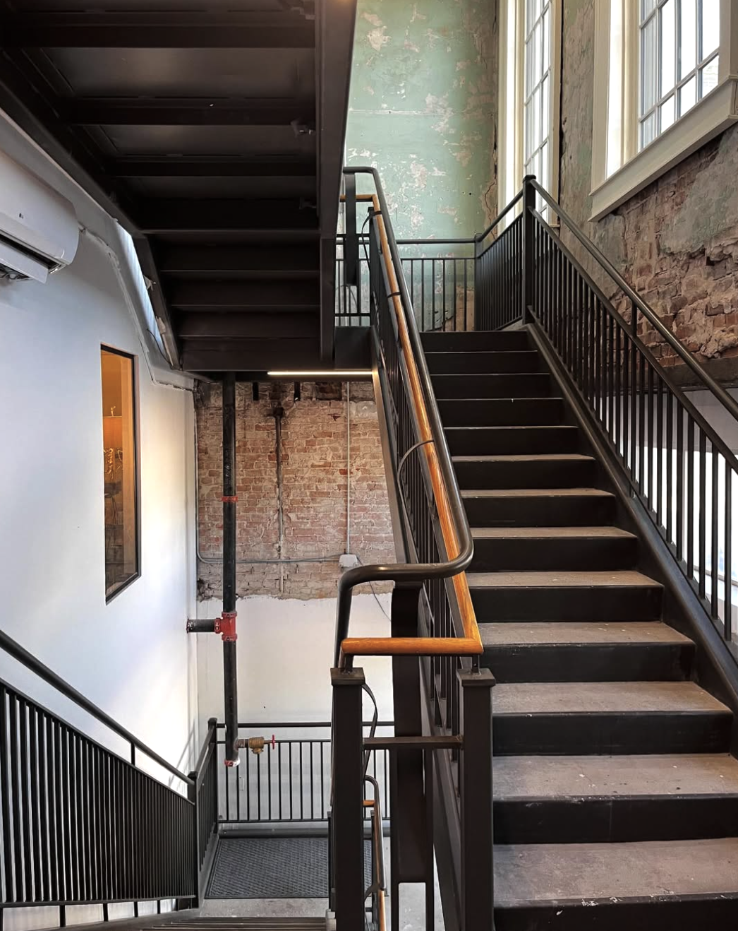 Interior of a building with black metal staircases and railings, an exposed brick wall, peeling green paint on the upper wall, large windows letting in natural light, and an air conditioning unit.