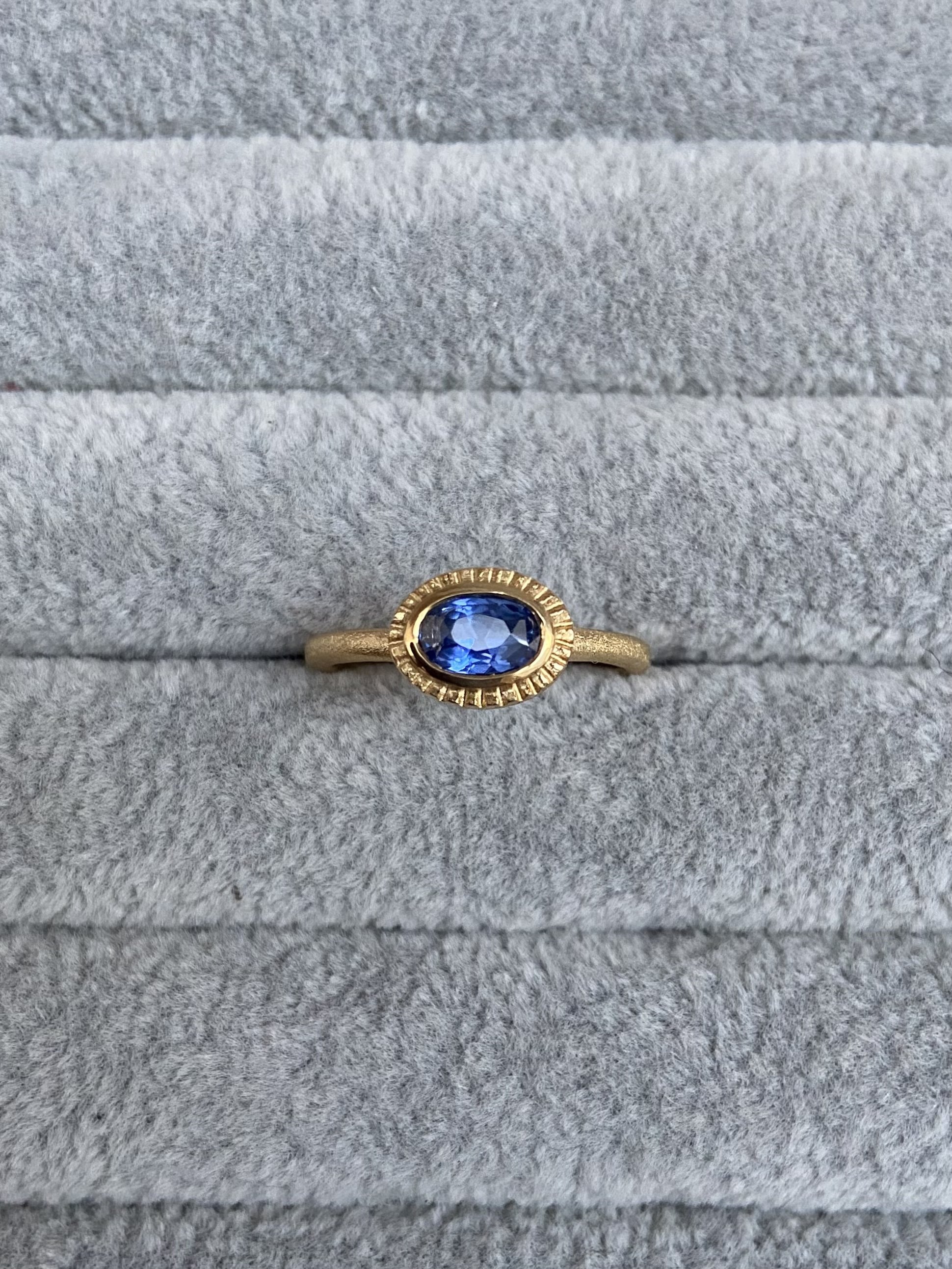 A gold ring with a large oval blue sapphire set in the centre, surrounded by a decorative gold border with small engraved details, resting on a velvet grey textured fabric surface.