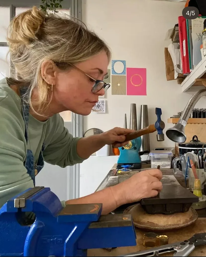 A woman wearing glasses and a green sweatshirt is working at a jeweller metalworking station, hammering metal on a work surface with tools and equipment around her.