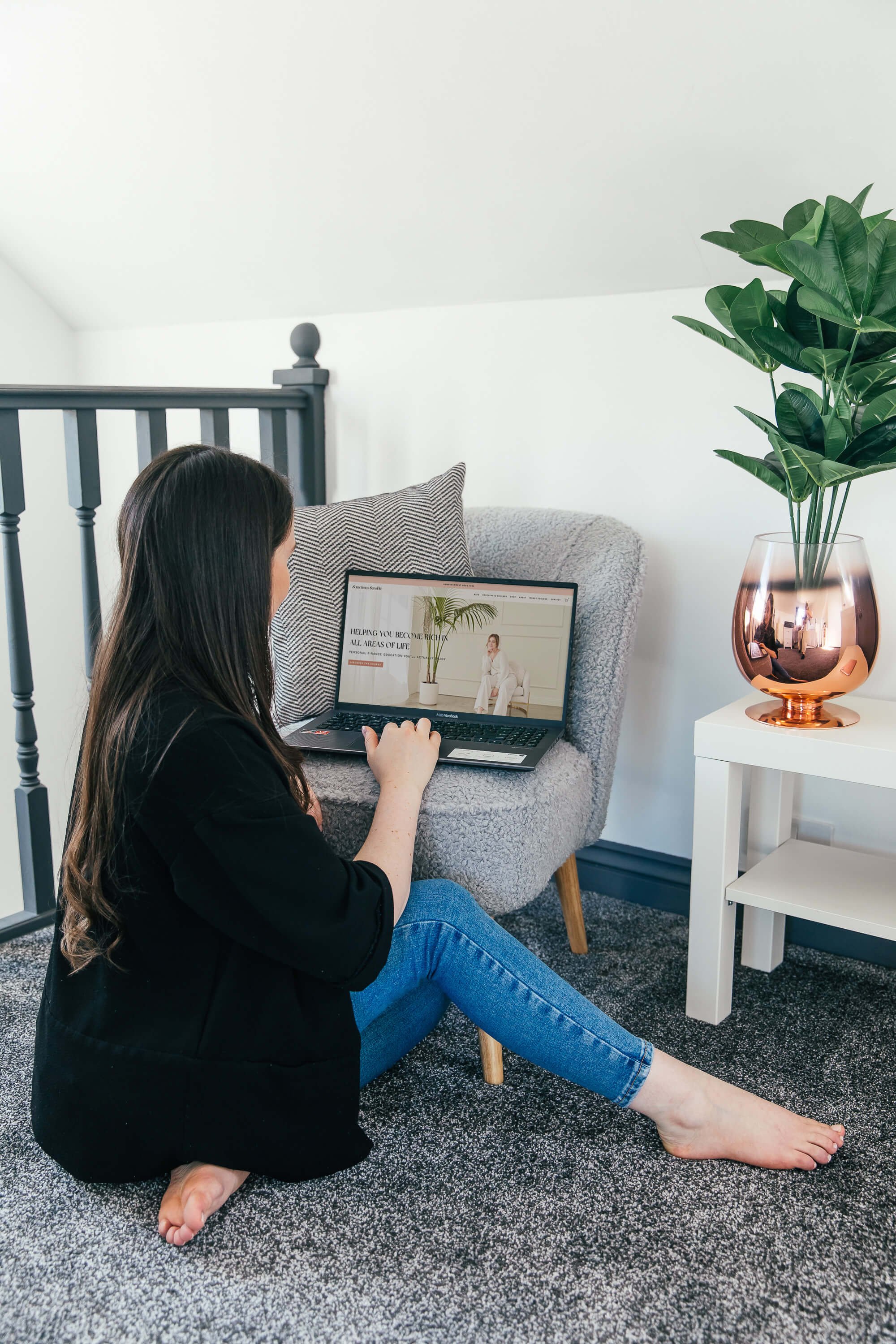 A woman sitting on the floor next to a grey armchair, working on a laptop placed on the chair. There is a large leafy plant in a copper-colored pot on a white side table beside her, and a black and white pillow on the armchair.