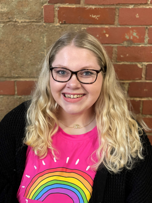 A woman with long blonde curly hair and glasses smiling at the camera, wearing a pink shirt with a rainbow graphic and a black cardigan, standing in front of a brick wall.
