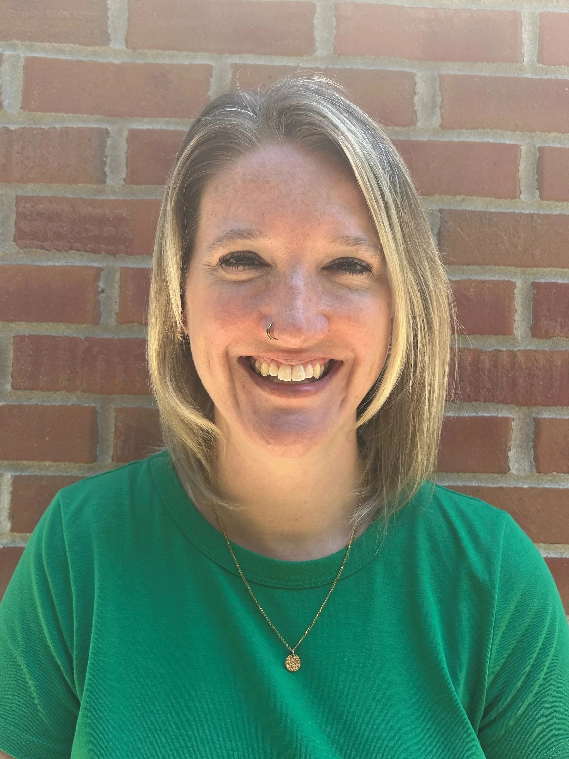 A woman with shoulder-length blonde hair, wearing a green shirt and a necklace, smiling in front of a brick wall.